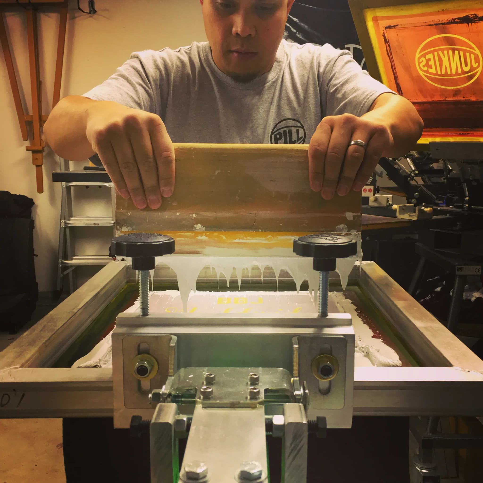 A printer pulling a flood bar of white ink across a screen clamped in the press, hands either side of the squeegee, an orange JUNKIES neon burning on the wall behind him.