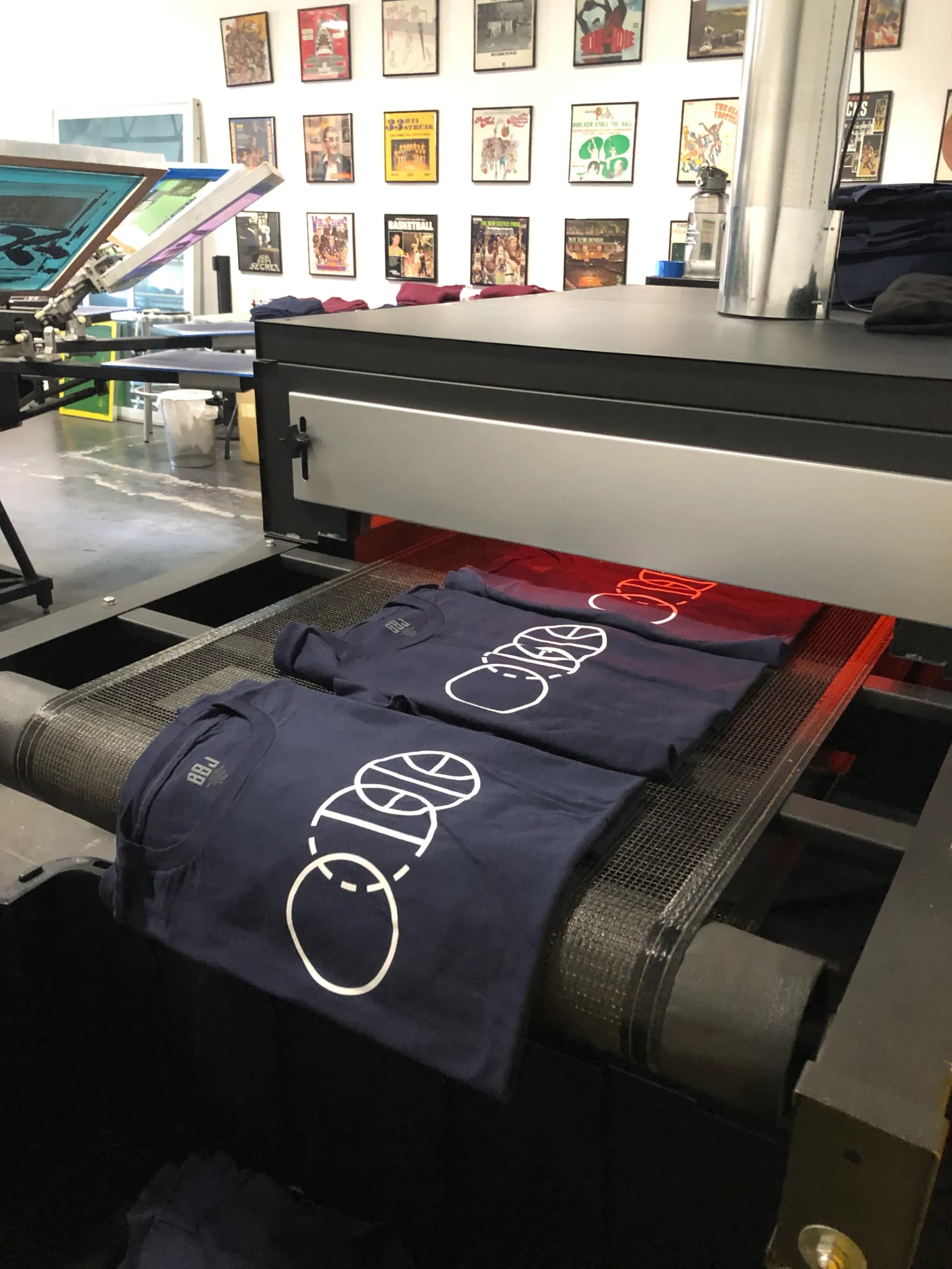 Three navy tees printed with the BBJ three-ring icon in white riding the mesh belt out of the conveyor dryer, a red print glowing under the heat behind them, the press and a wall of framed posters beyond.