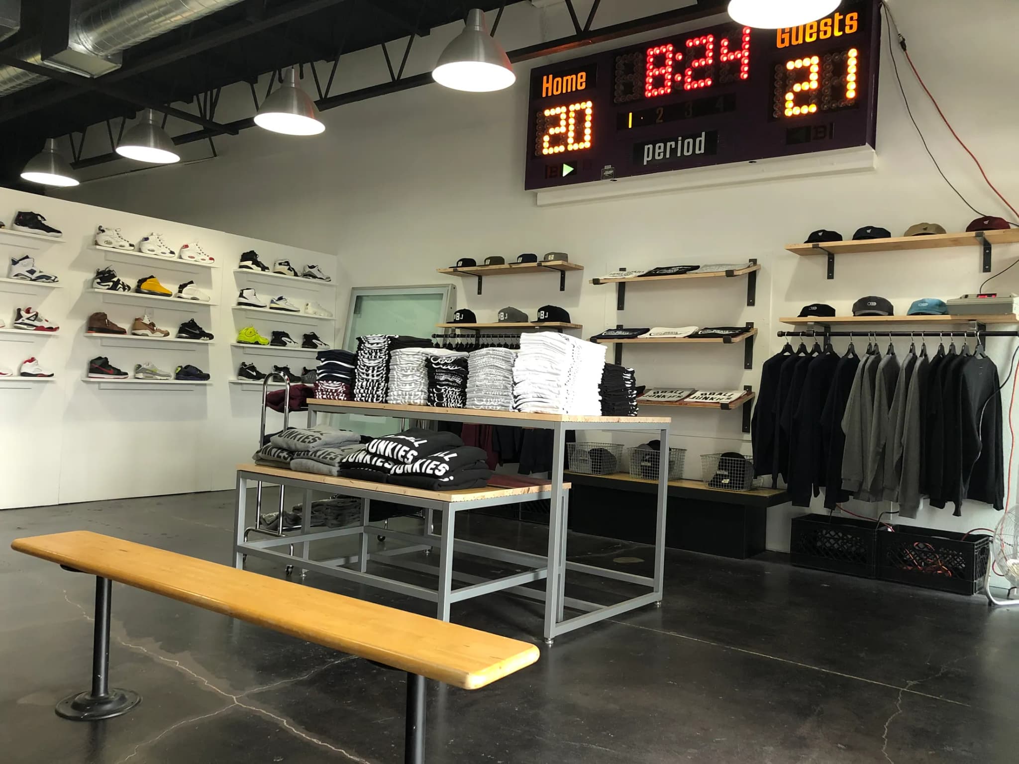 The shop floor under a working scoreboard: sneakers on wall shelves, caps and folded tees on a parquet-topped table, fleece on a rail, and a court bench across the foreground.