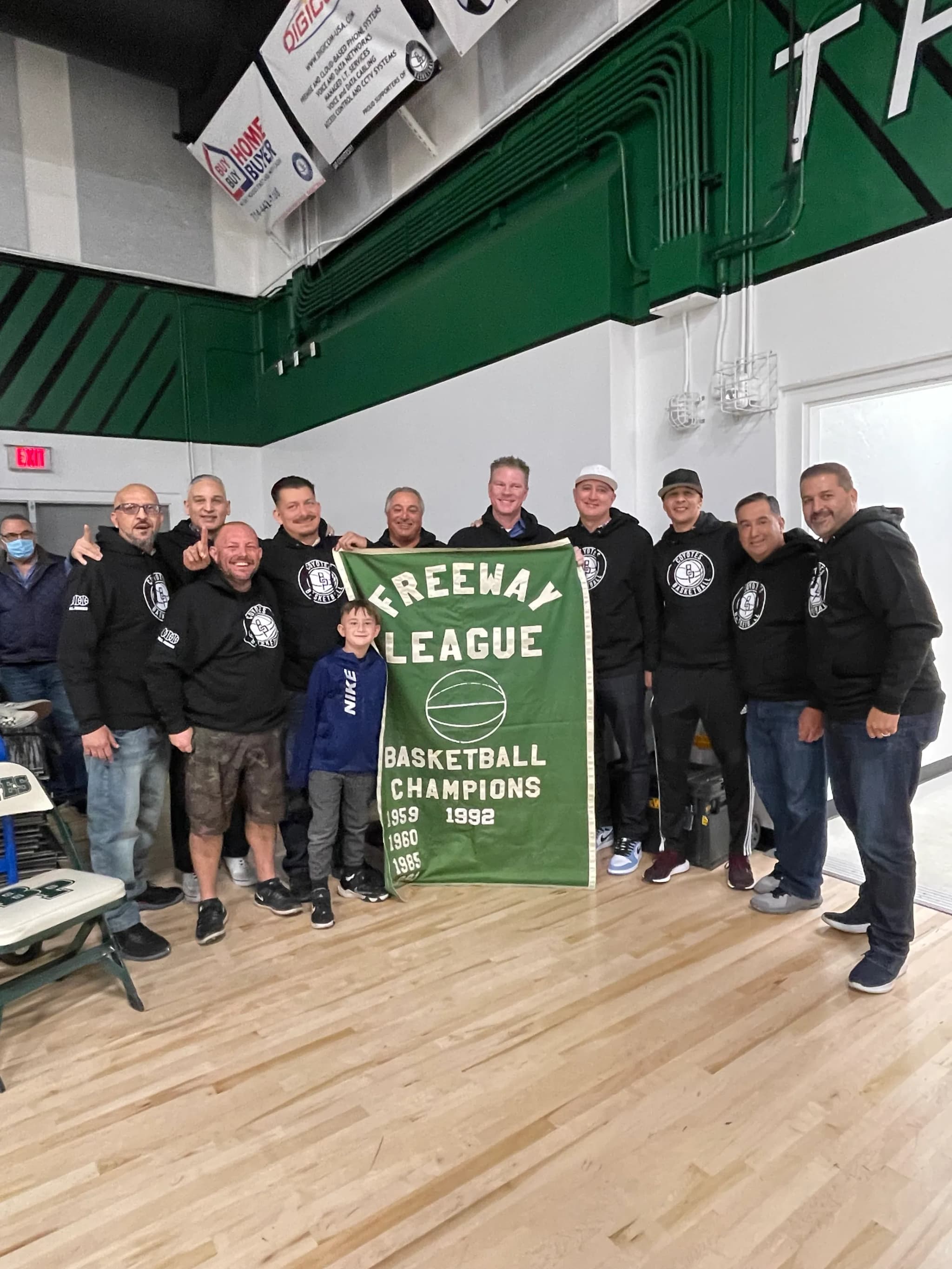 Eleven adults and a child in matching black hooded sweatshirts carrying a school basketball crest, holding open a green FREEWAY LEAGUE BASKETBALL CHAMPIONS banner on a gym floor.