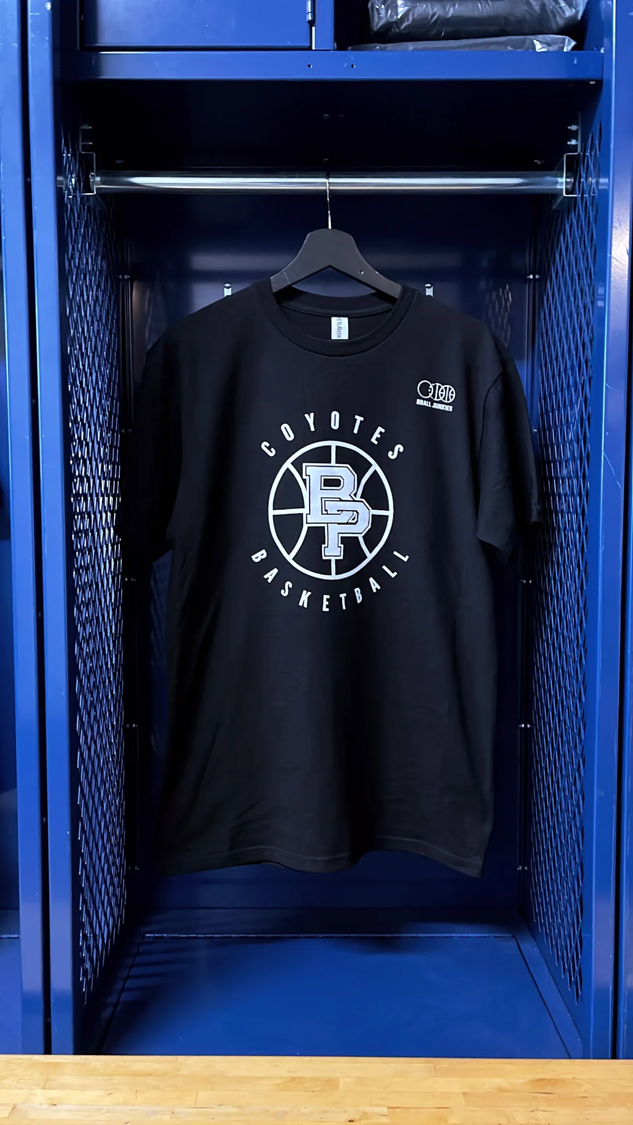 A navy tee on a wooden hanger inside a lit blue locker, screen printed at the chest with a school basketball roundel in white.