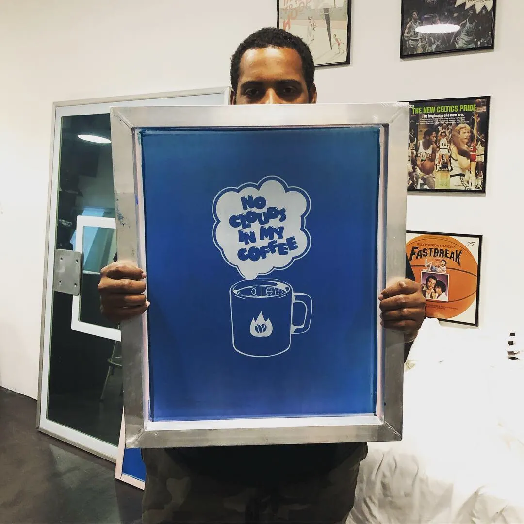 A man holding up a freshly burned screen in its aluminum frame, the emulsion carrying a steaming mug and a speech cloud reading NO CLOUDS IN MY COFFEE.