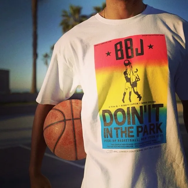 A natural tee held up at dusk with palms behind it, printed with a sunset-gradient poster panel for the film over a silhouetted layup and the BBJ wordmark, a ball tucked under the wearer’s arm.