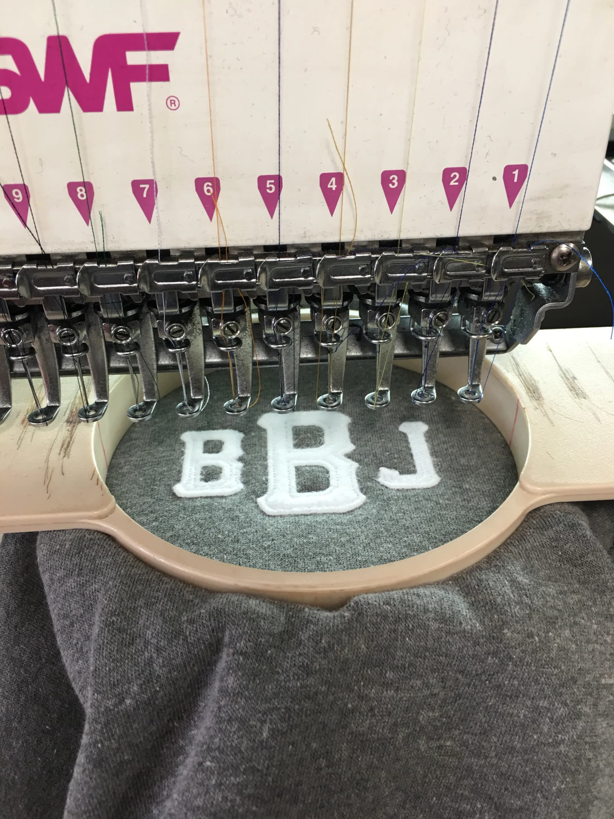 A multi-head embroidery machine with a garment hooped under the needle bar, BBJ appliqué letters stitched into gray fleece beside a numbered thread guide.