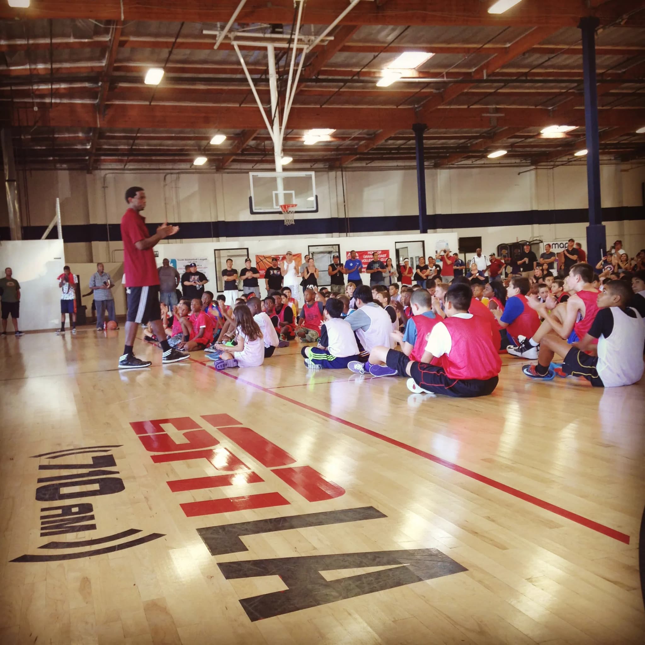 A coach addressing rows of kids seated on a gym floor at a youth clinic, the court painted with a broadcaster’s marks under them.