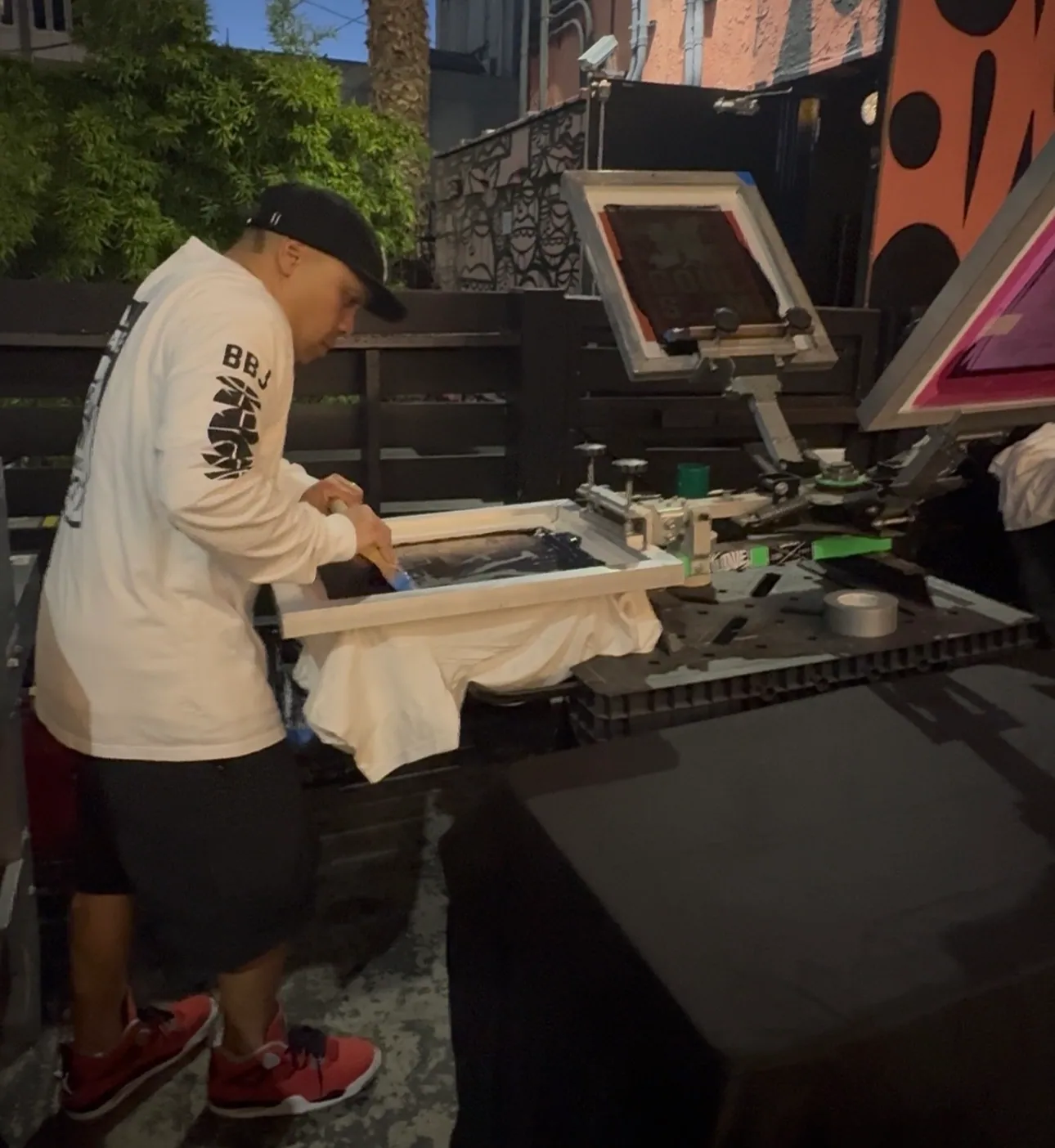 A printer in a BBJ tee bearing down on a squeegee across a garment on a manual press set up outdoors, a rack of screens behind him.