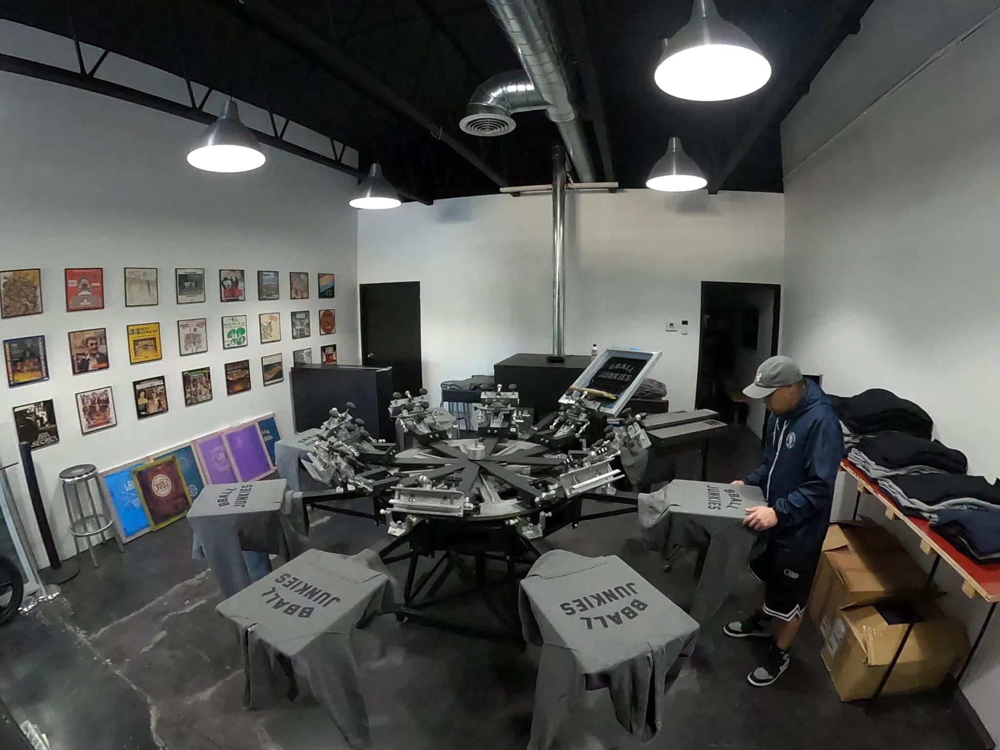 The print floor from the doorway: a six-station carousel press in the middle of the room, garments laid out on two tables and framed artwork covering the far wall.