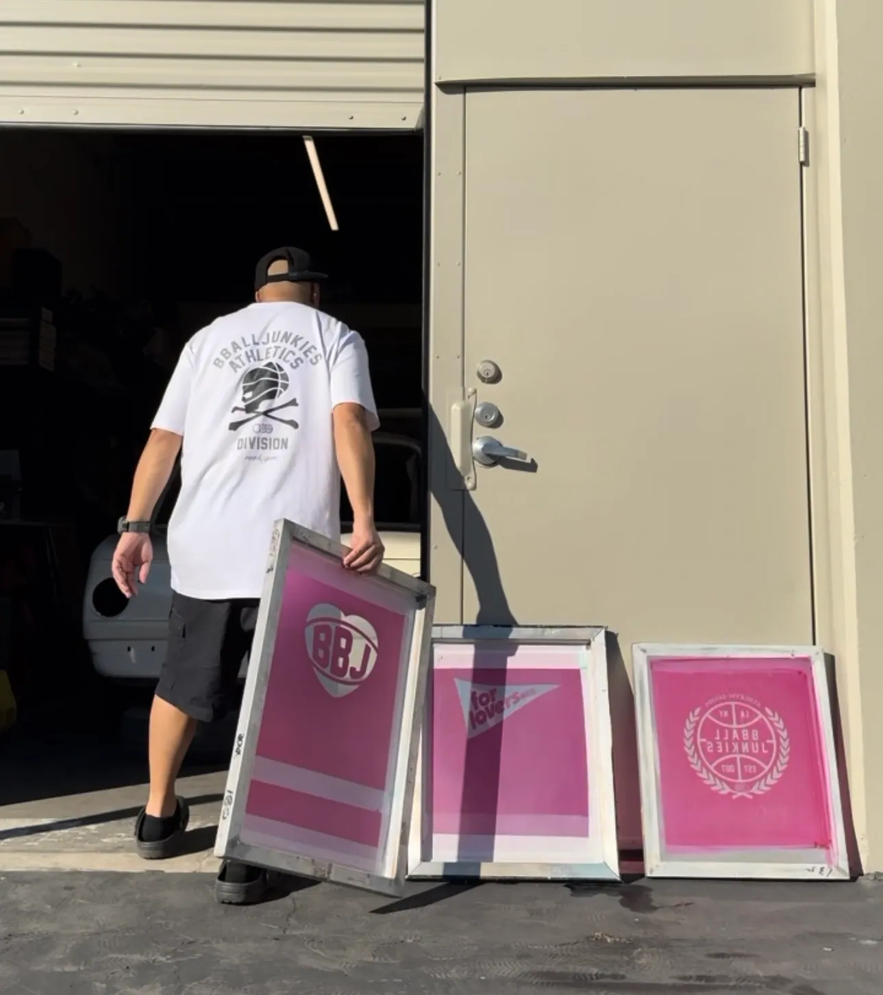 A printer carrying three burned screens in pink emulsion through the shop’s roll-up door, a BBJ mark reading in reverse on each one.