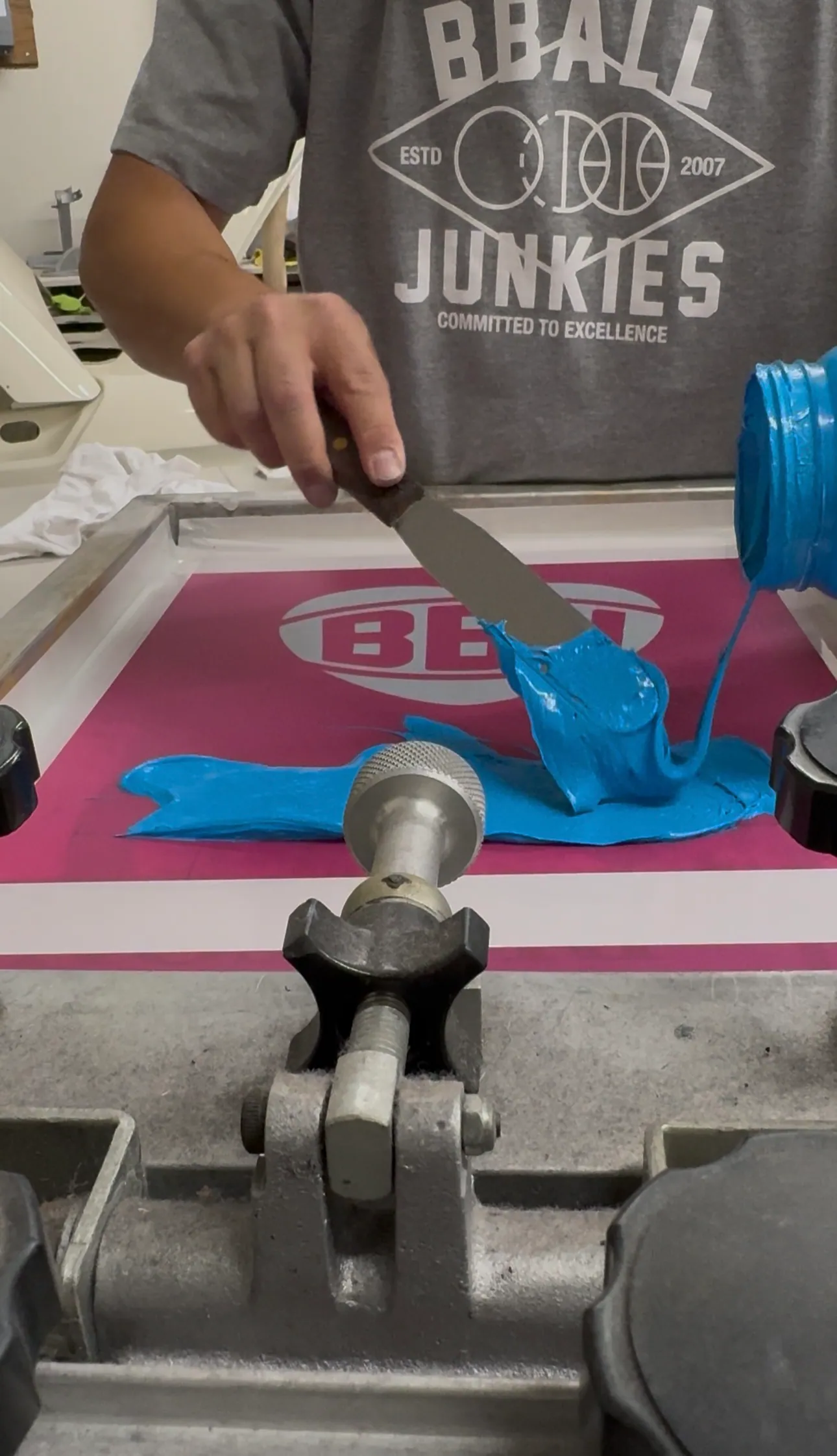 A hand pouring blue ink from a jar onto a screen clamped in the press, a BBJ wordmark stencilled in magenta emulsion beneath it.