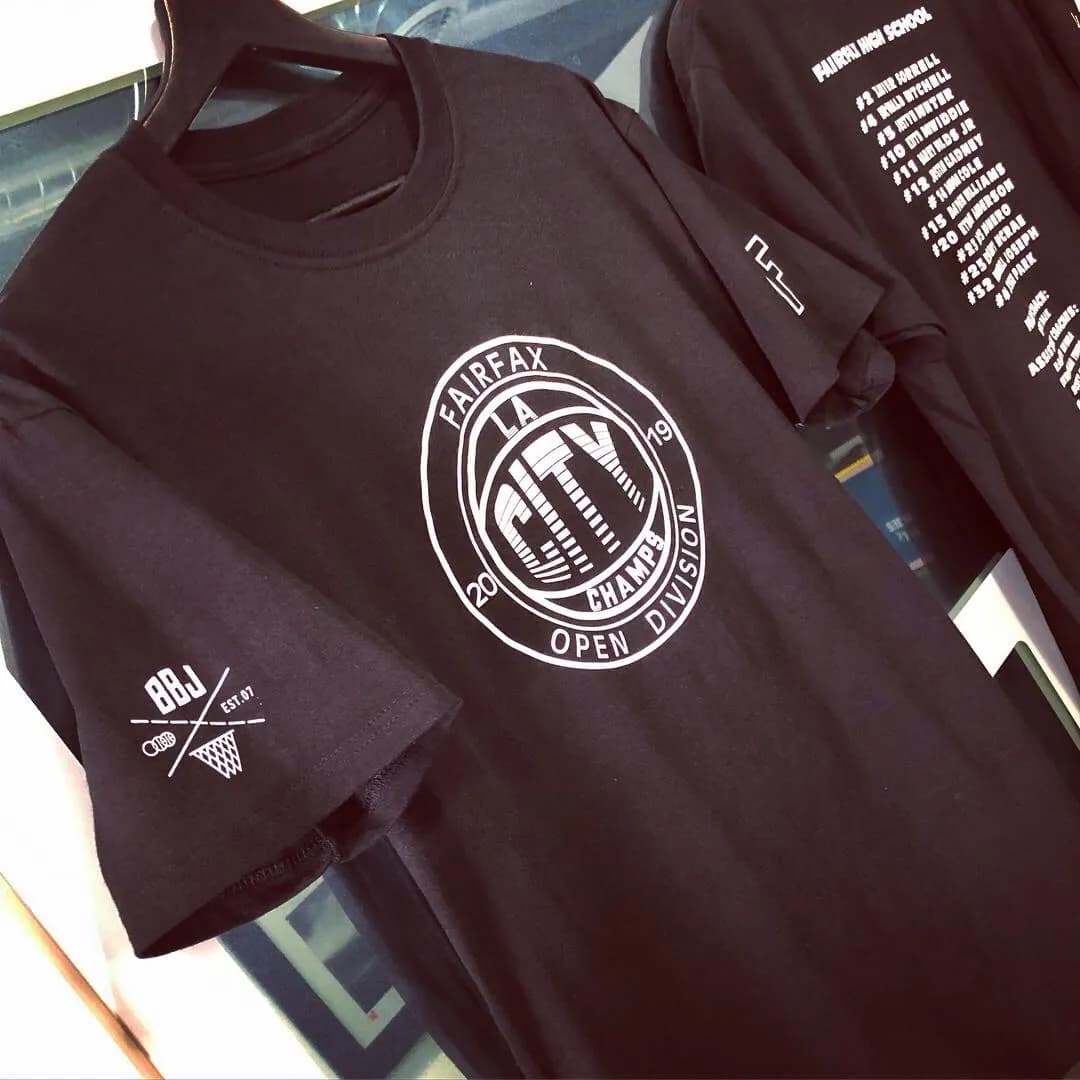 A maroon tee printed with a Fairfax High School championship crest at the chest and the BBJ crossed-line mark small beside it, folded on a stack.