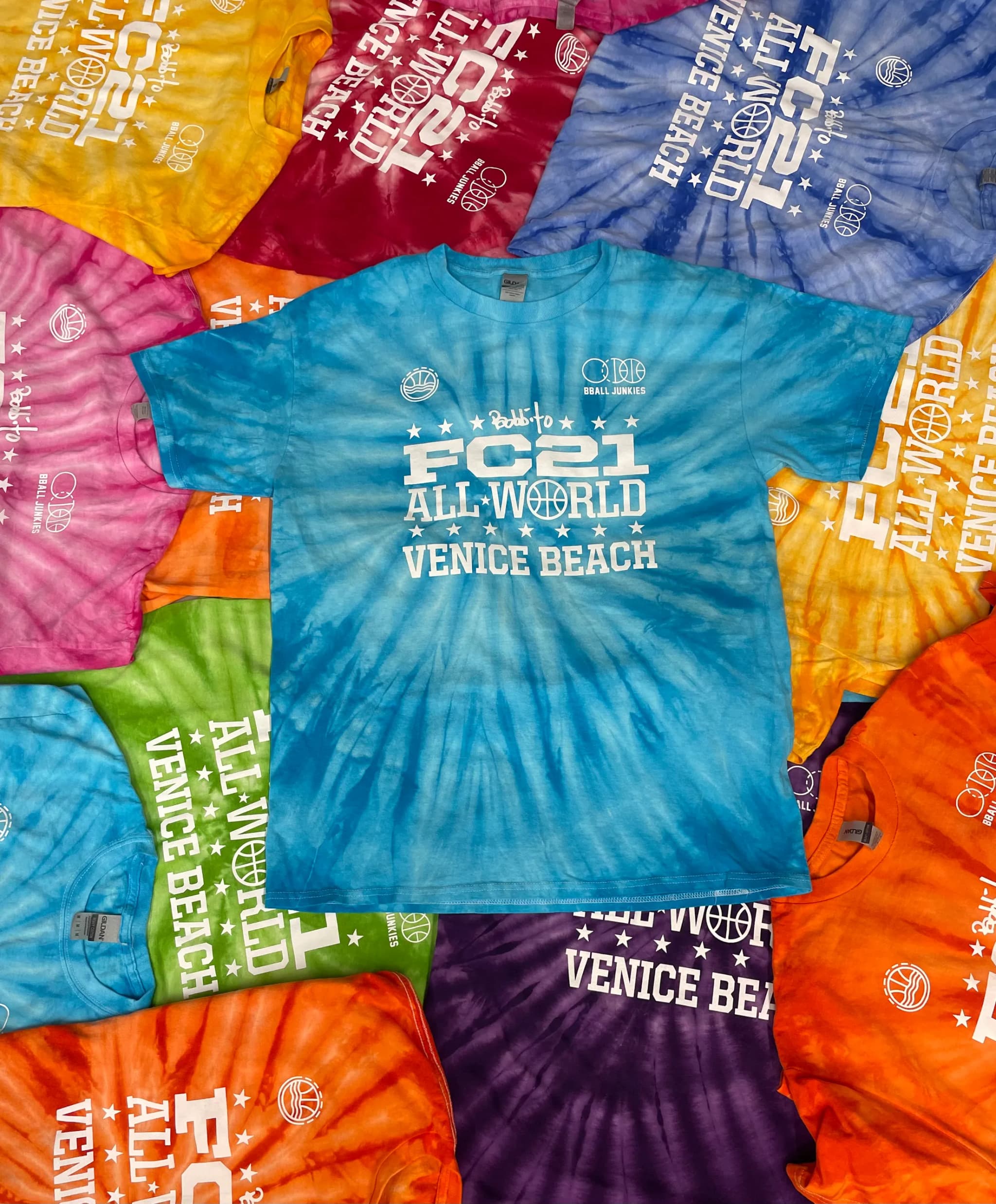 A fan of tie-dyed tournament tees in a dozen colorways, each printed FC21 ALL-WORLD · VENICE BEACH, laid out overlapping with a light blue one on top.