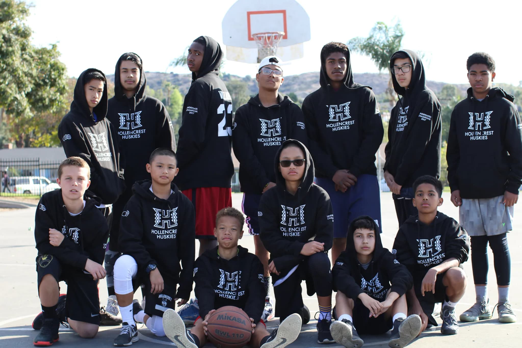A middle-school squad on an outdoor court in matching black hooded sweatshirts carrying the school's monogram, ranged under the hoop with a ball at their feet.