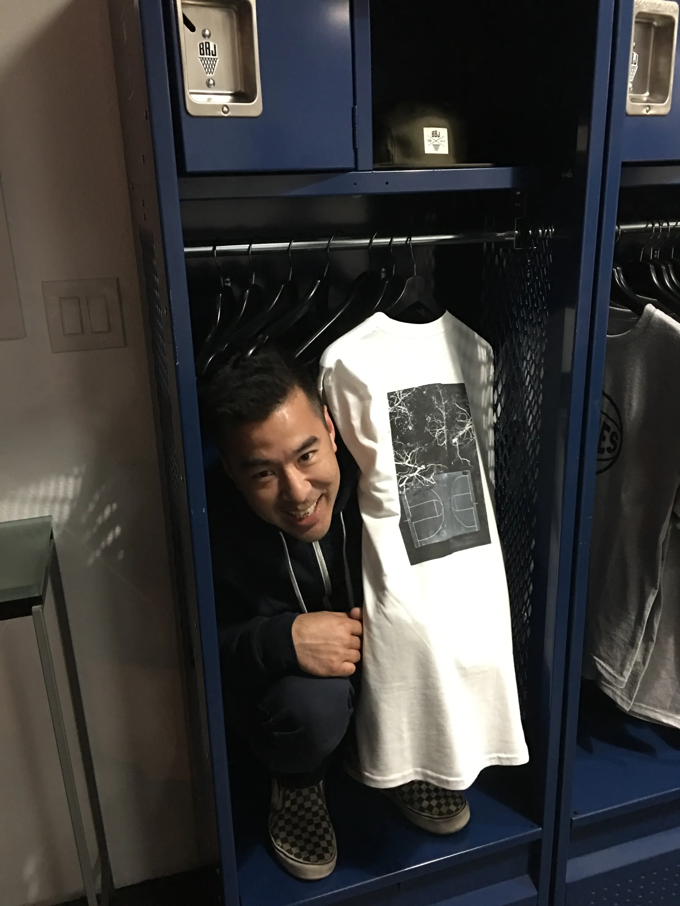 A photographer in a printed tee and black cap standing in front of a bank of blue lockers in the shop, holding a rolled garment.