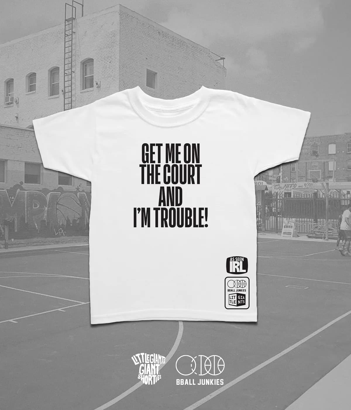 A white youth tee laid over a photograph of an outdoor city court, printed GET ME ON THE COURT AND I’M TROUBLE! in four stacked lines, with an AS SEEN IRL stamp and a co-signed BBALL JUNKIES and LITTLE GIANTS box at the hem.