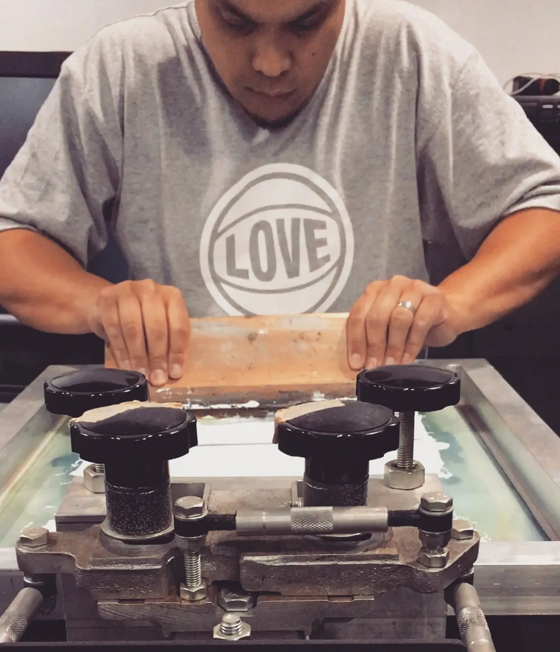 A printer in a BBJ LOVE tee bearing down on a squeegee across a screen clamped in a portable press, the flood stroke half pulled.