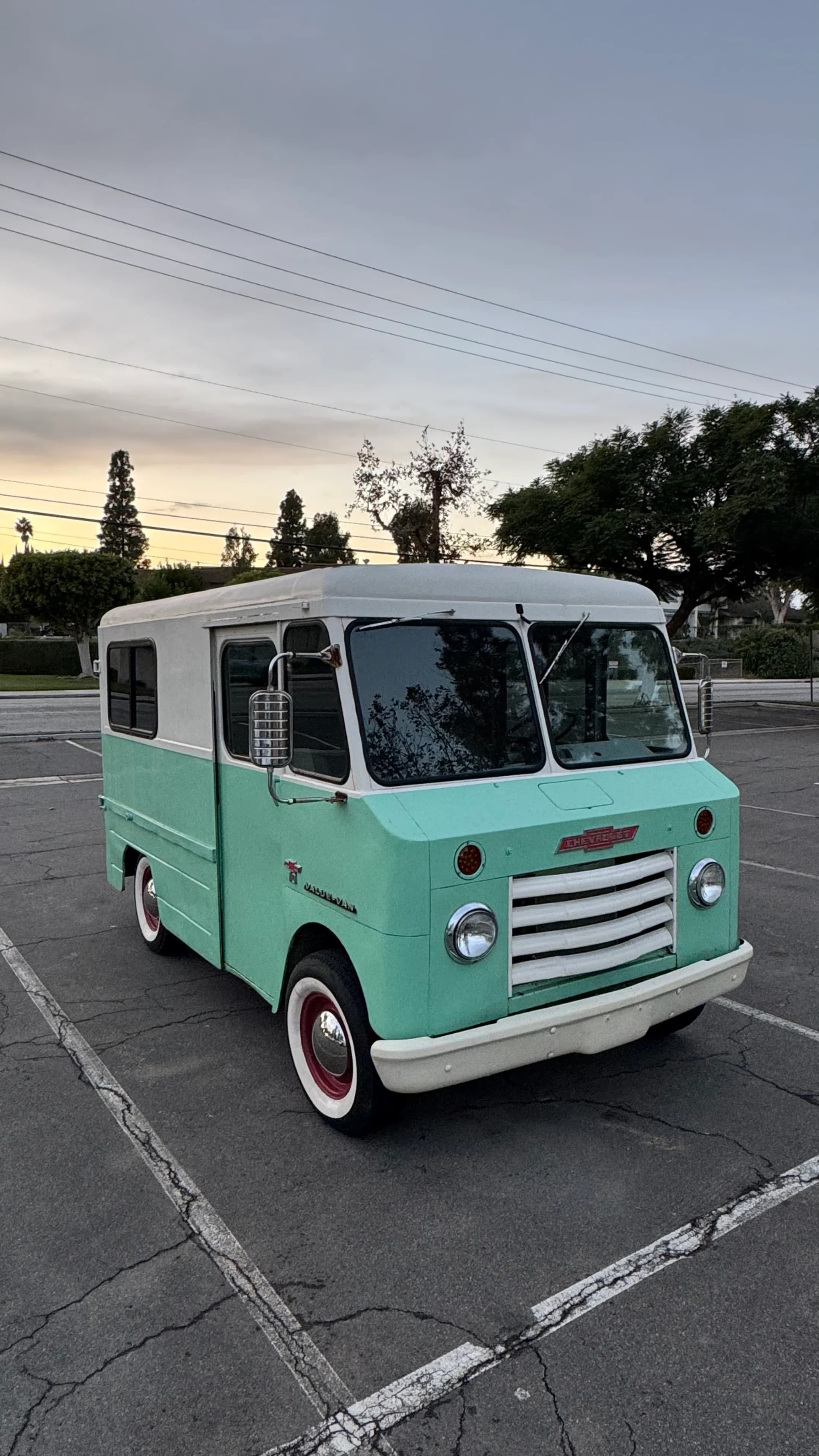 A restored step van in two-tone seafoam green and white, parked at dusk with its chrome grille and whitewall tires catching the last light.