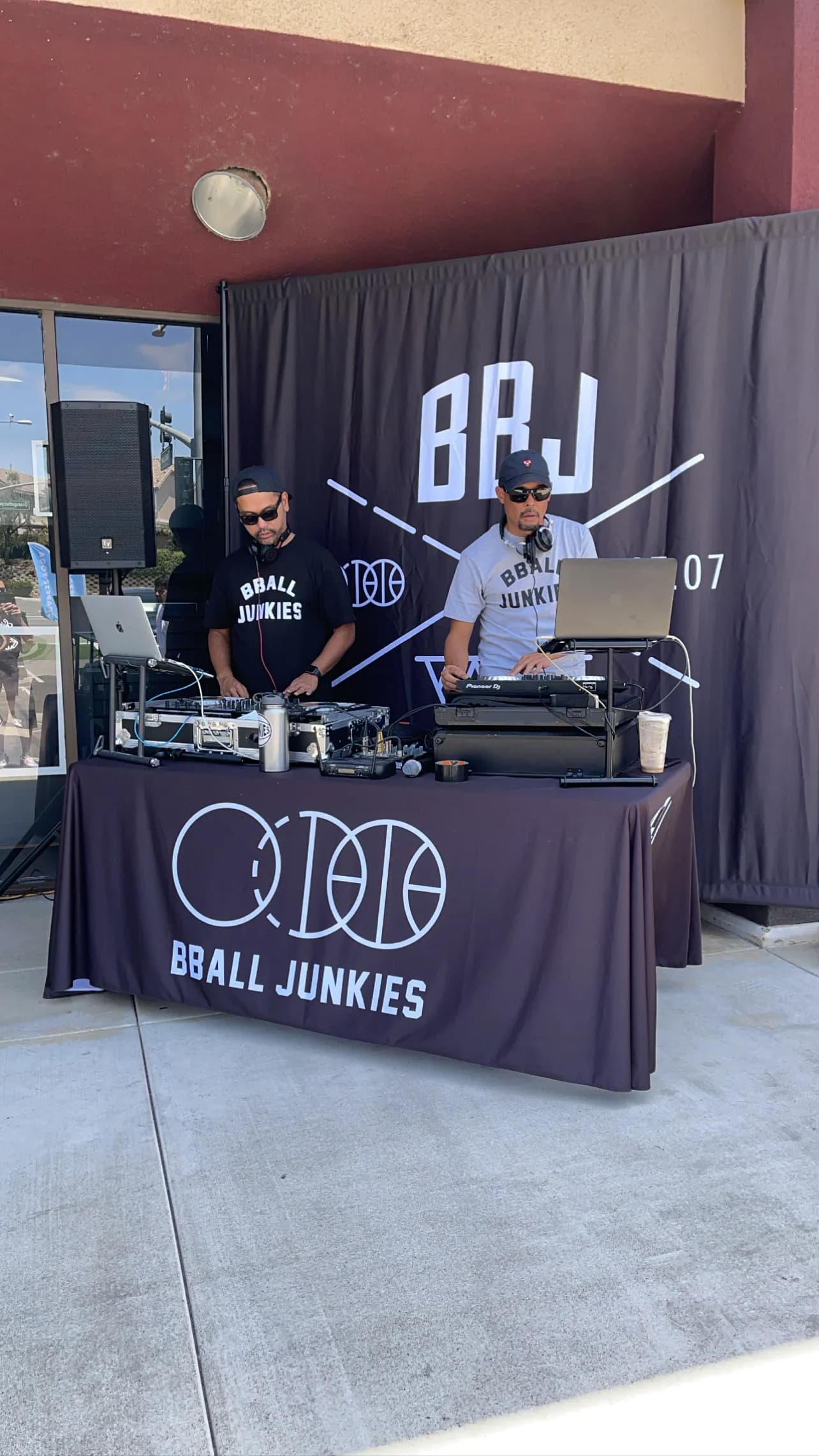 Two DJs behind a draped BBALL JUNKIES table at an event, a BBJ backdrop banner and a PA stack behind them.