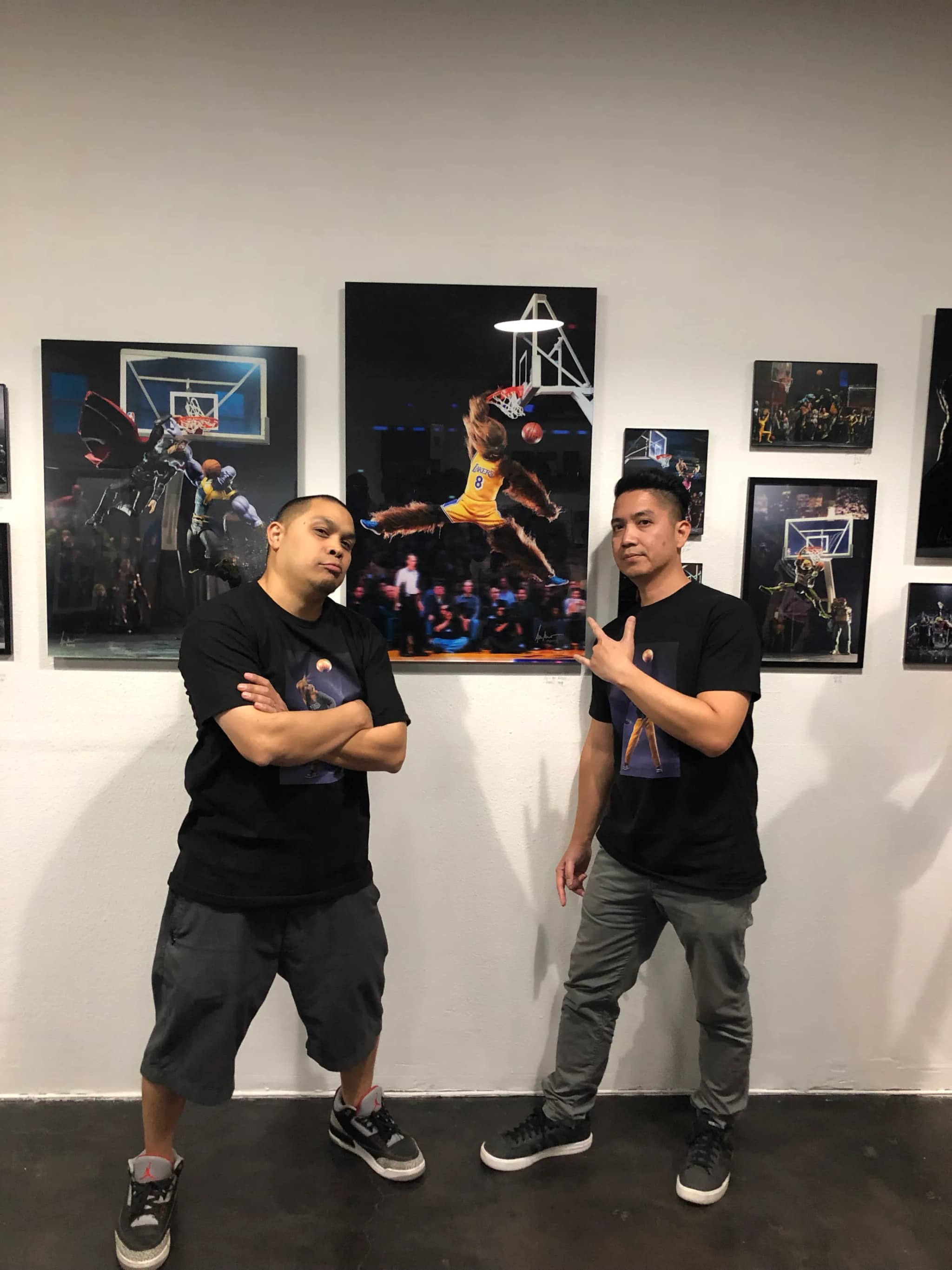 Two people in black standing at a gallery wall of framed action-figure photographs during the opening, the shop’s painted court floor under them.