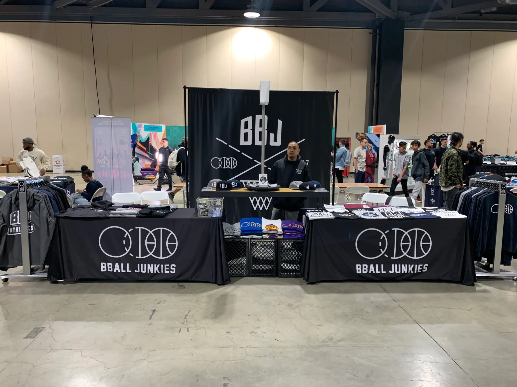 The pop-up booth set up indoors: two draped BBALL JUNKIES tables of folded tees and caps under a printed backdrop banner, with a rack of garments behind.