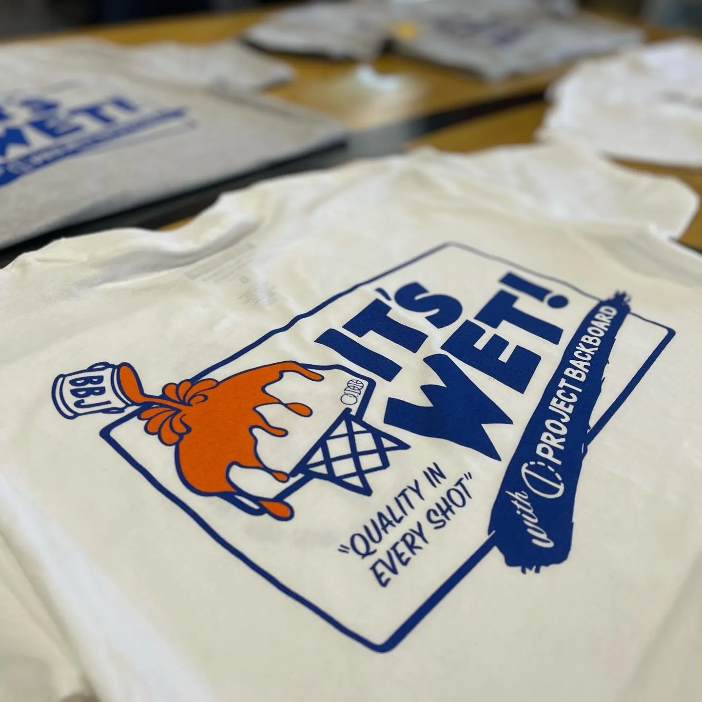 A natural cotton tee folded on a table, screen printed in blue and orange with a hand dunking a dripping ball through a net over the words IT’S WET! and "Quality in every shot", co-signed BBJ and Project Backboard.