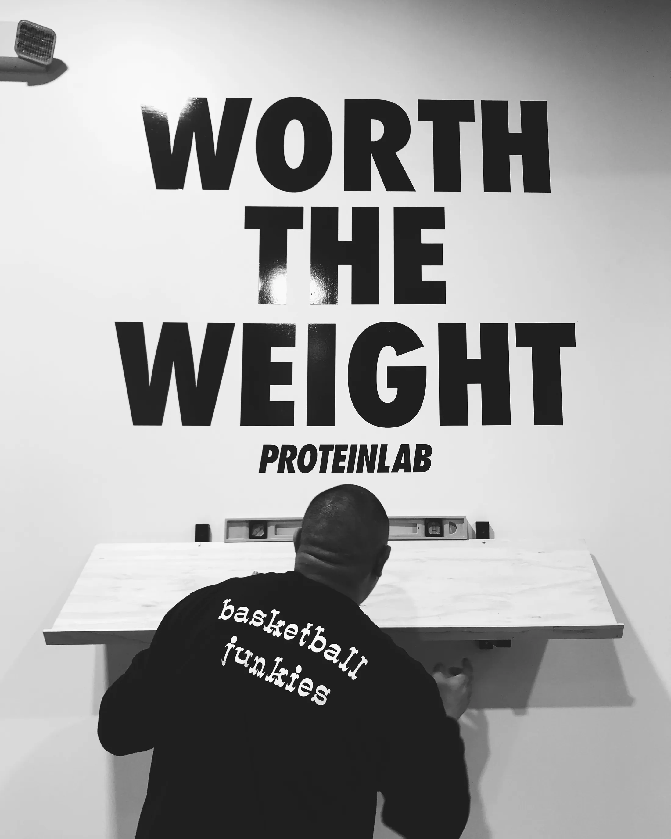 A man in a hooded sweatshirt lettered "basketball junkies" levelling a white counter beneath a wall painted WORTH THE WEIGHT · PROTEINLAB.