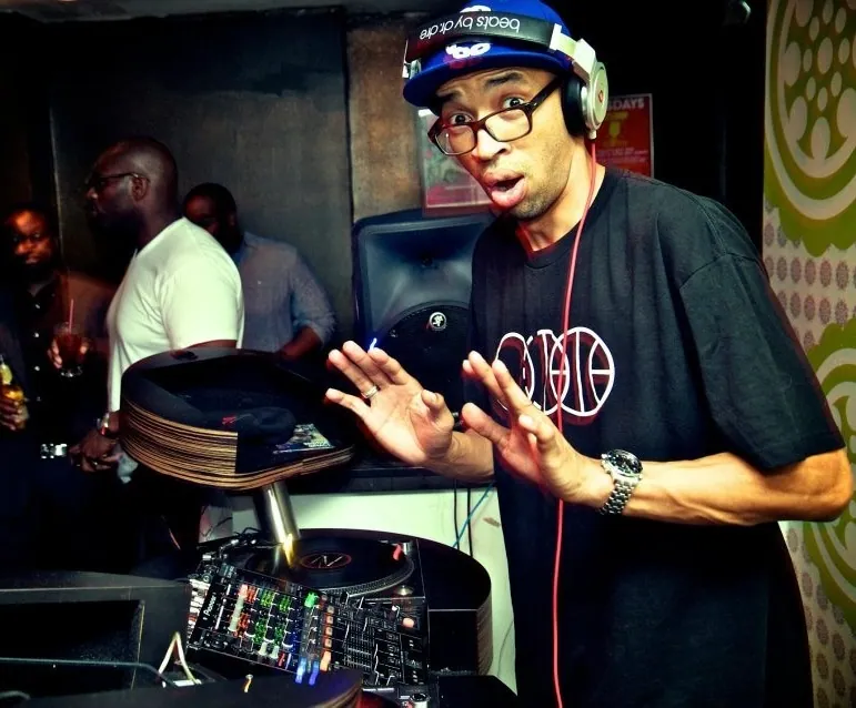 A DJ in a BBJ three-ring tee working a pair of turntables at a club night, headphones on, one hand raised off the platter.