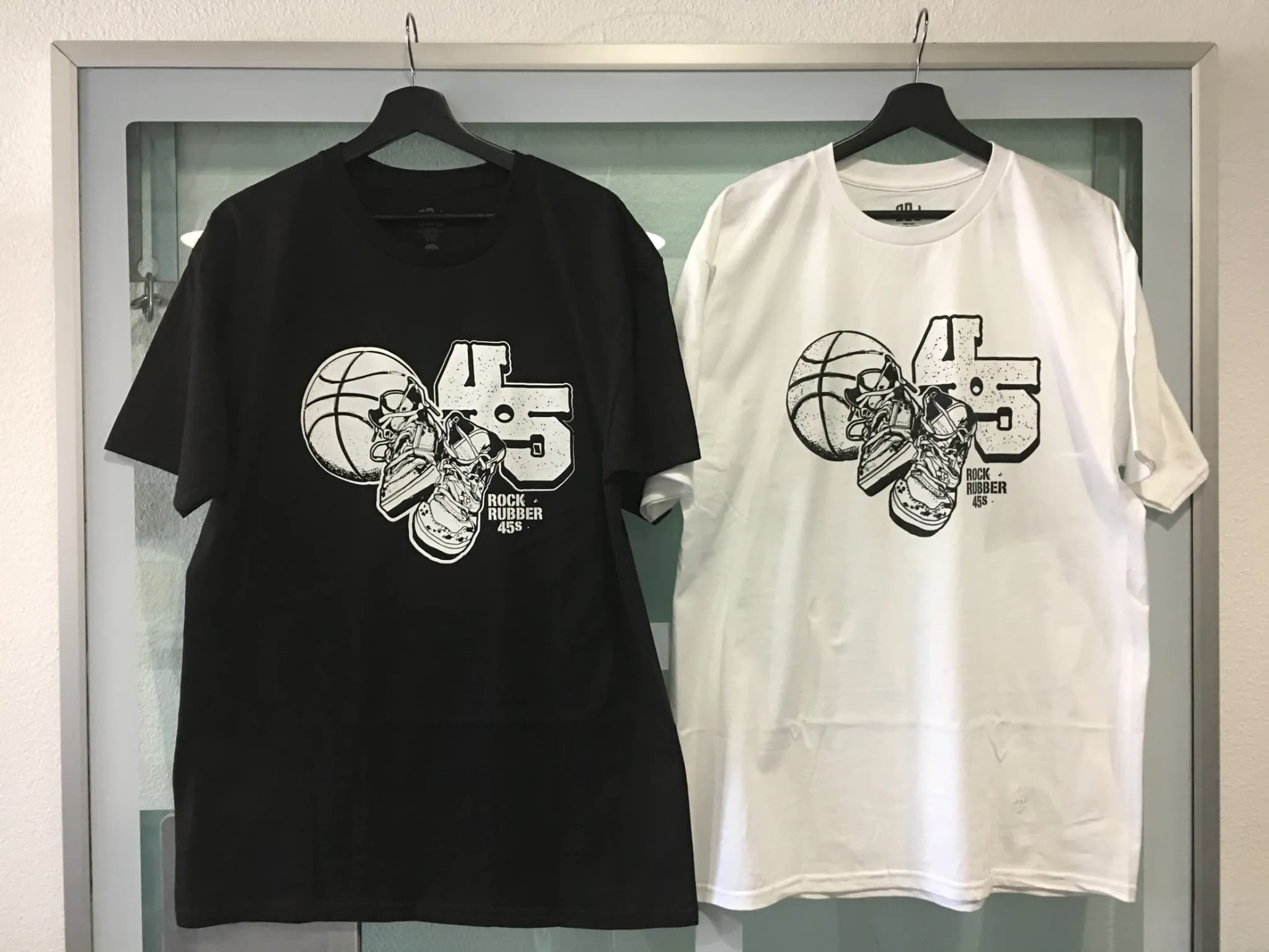 A black tee and a white tee on hangers side by side in a display case, each printed with a 45 record, a ball and a sneaker locked together as one mark over ROCK RUBBER 45s.