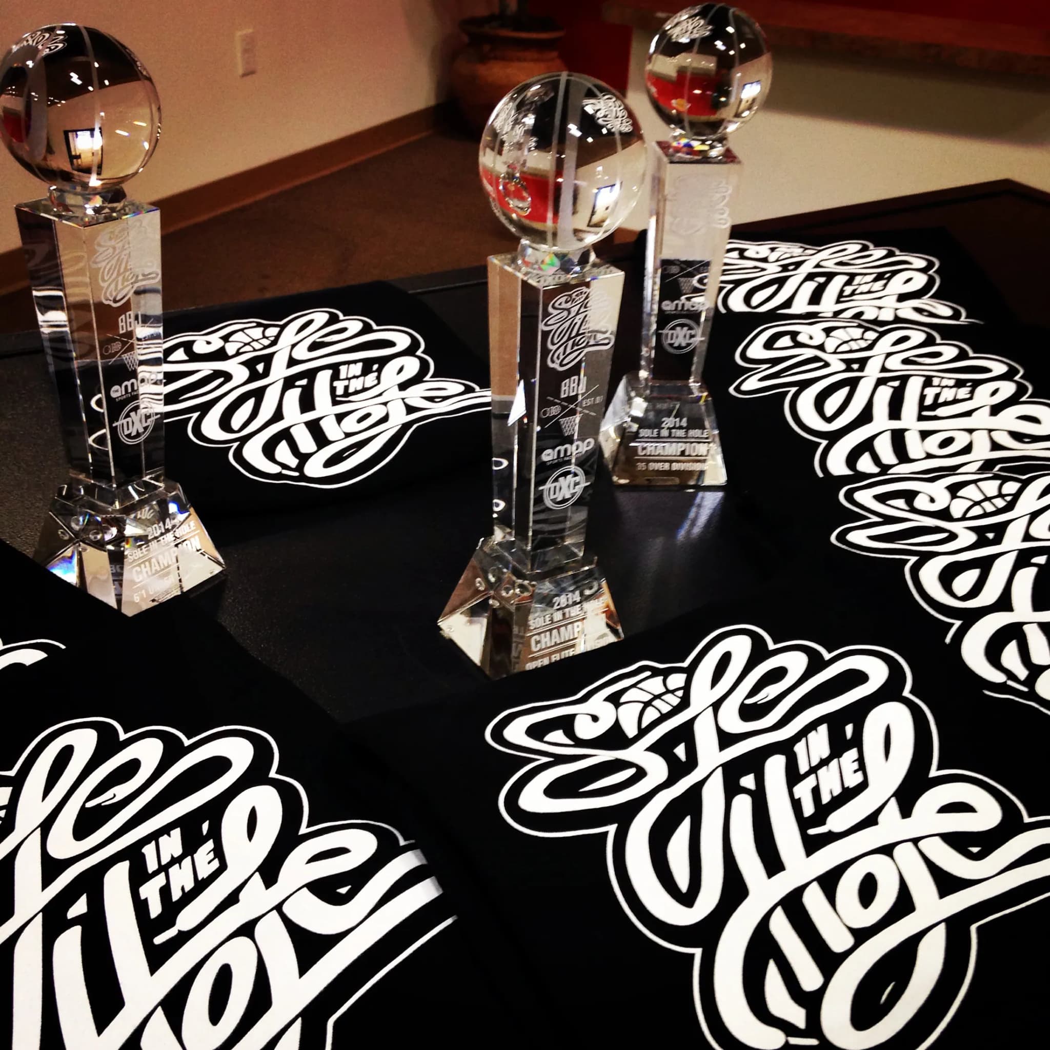 Three glass tournament trophies with basketballs set in their tops, standing on a table spread with black championship tees lettered SOLE IN THE HOLE in script.