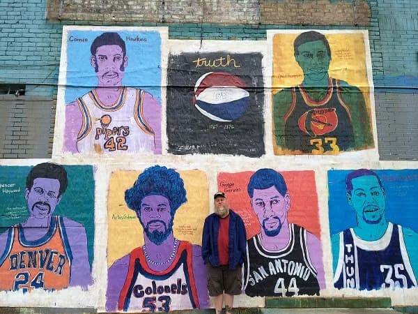A painted brick wall of portrait panels — basketball players of several eras in their team colors, a ball and a hand-lettered TRUTH panel among them — with a man standing in front of it.