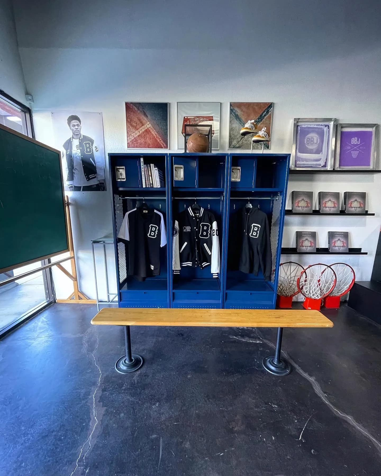 The three blue steel lockers pulled back to show the chalkboard beside them, a B jersey, a varsity jacket and a hoodie on the rails, framed court photographs above, a court bench in front and three netted hoops standing on the floor.