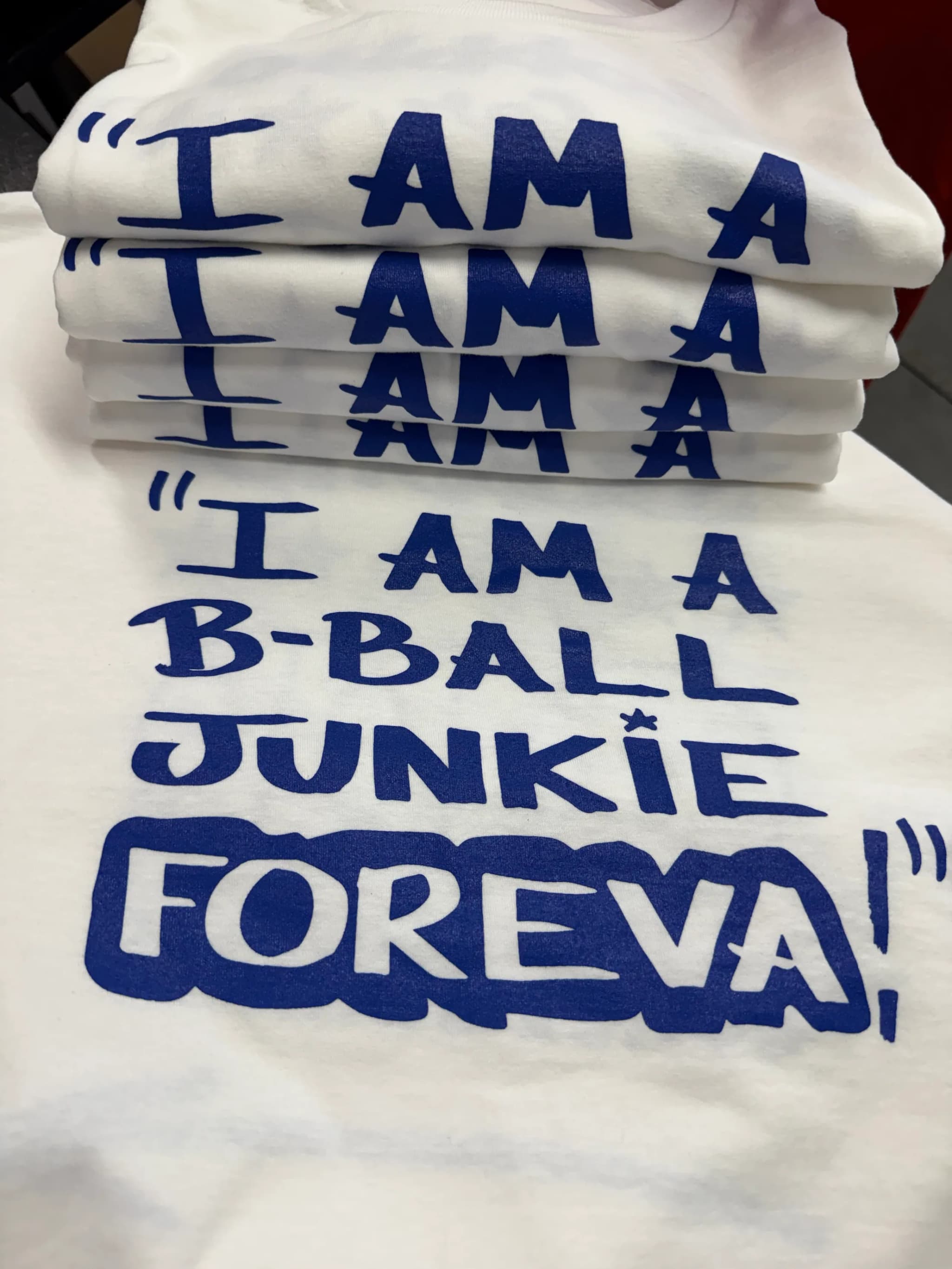 A stack of folded white tees, each printed in blue hand-lettering, the top one opened out to read I AM A B-BALL JUNKIE FOREVA! in full with the last word boxed.