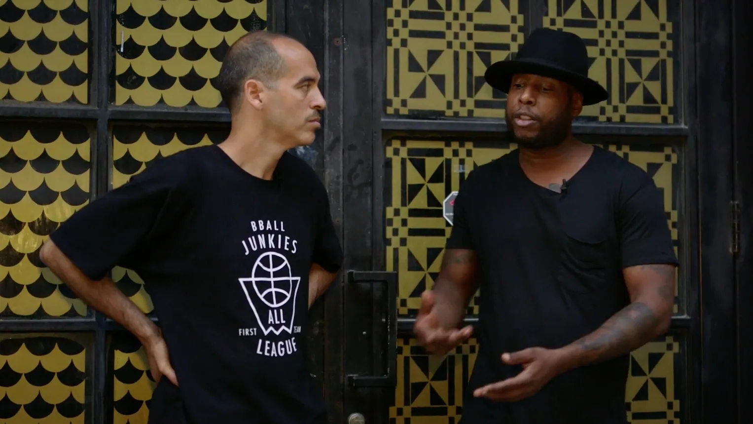 Two men talking outside a pair of tall doors patterned in gold and black, the one on the left in a black BBALL JUNKIES ALL LEAGUE tee with his hands on his hips, the one on the right in a wide-brimmed black hat gesturing as he speaks.