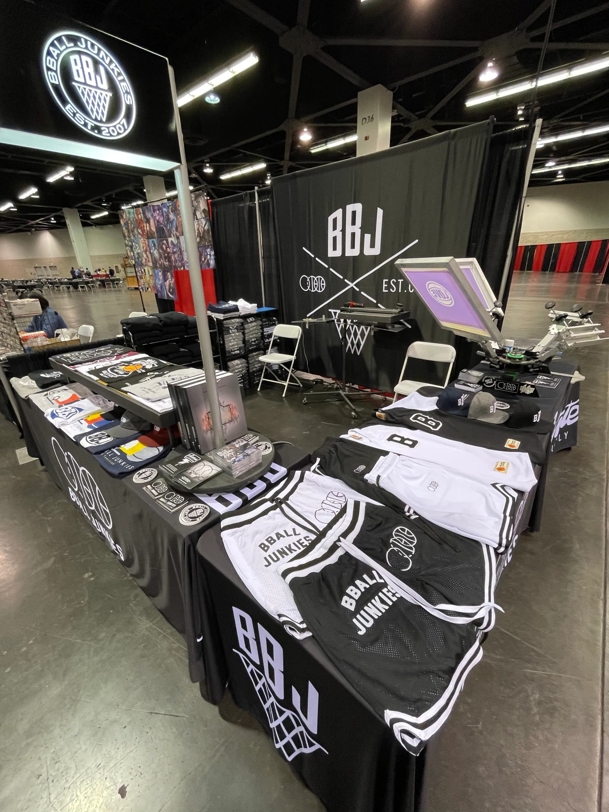 The BBJ booth on a convention floor: a lit BBALL JUNKIES roundel over the header, the crossed-line mark on a black backdrop, mesh jerseys and shorts laid across black-draped tables beside caps and folded tees, and a screen printing press racked at the end of the row.