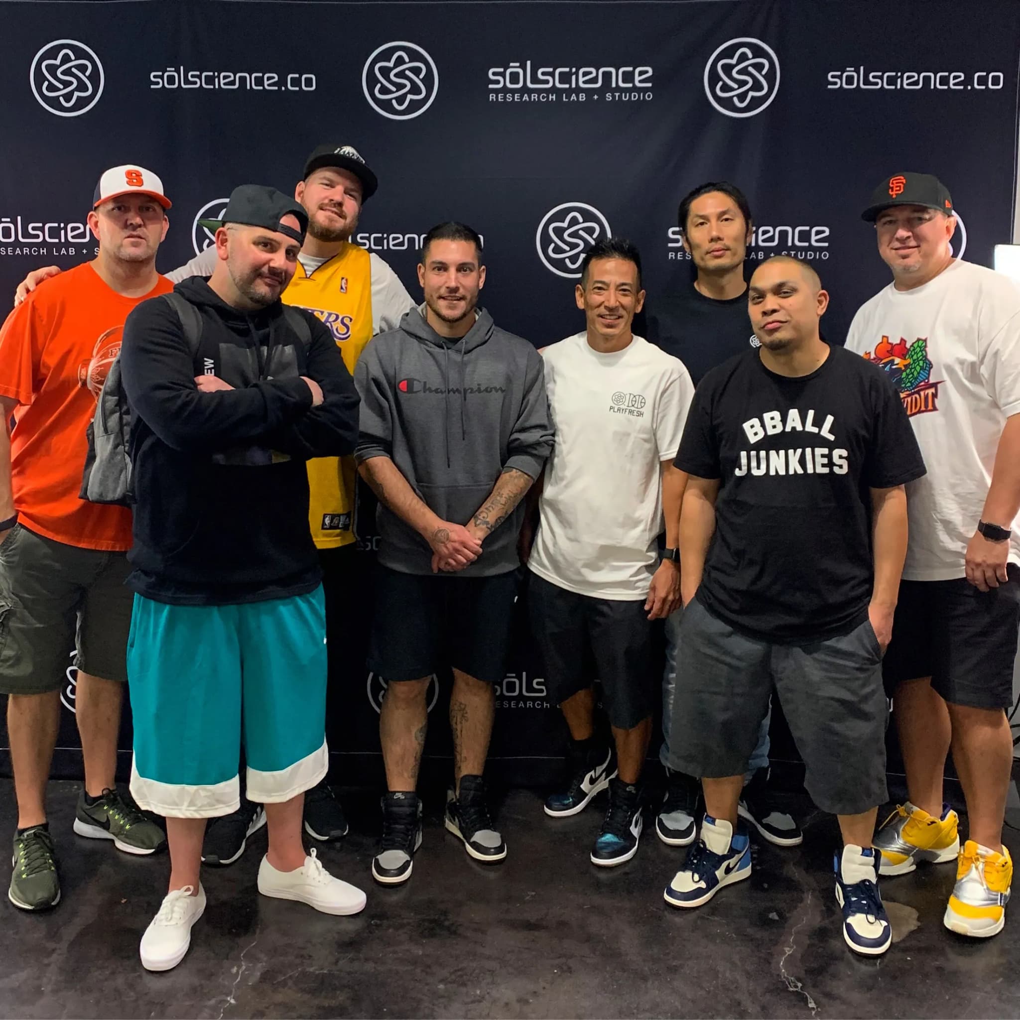 Eight men shoulder to shoulder in front of a sōlscience.co step-and-repeat banner, a ninth cut off at the left edge, one in a black BBALL JUNKIES tee and another in a Lakers jersey, every one of them in sneakers.