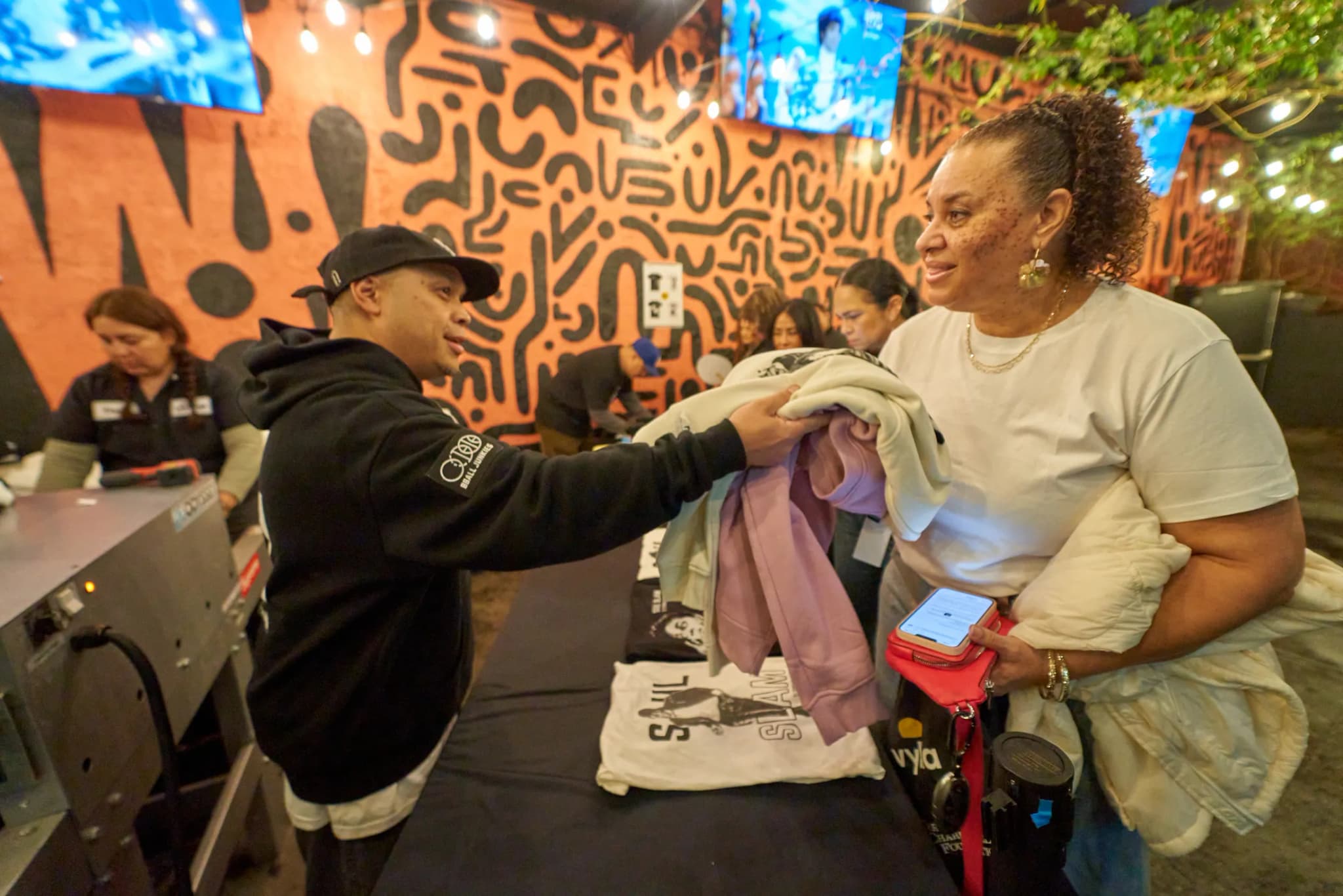 A printer in a black BBJ hoodie and cap handing a stack of freshly printed tees and sweatshirts across the merch table to a guest at the Echoplex, a Soul Slam tee laid out between them and an orange mural patterned in black behind.