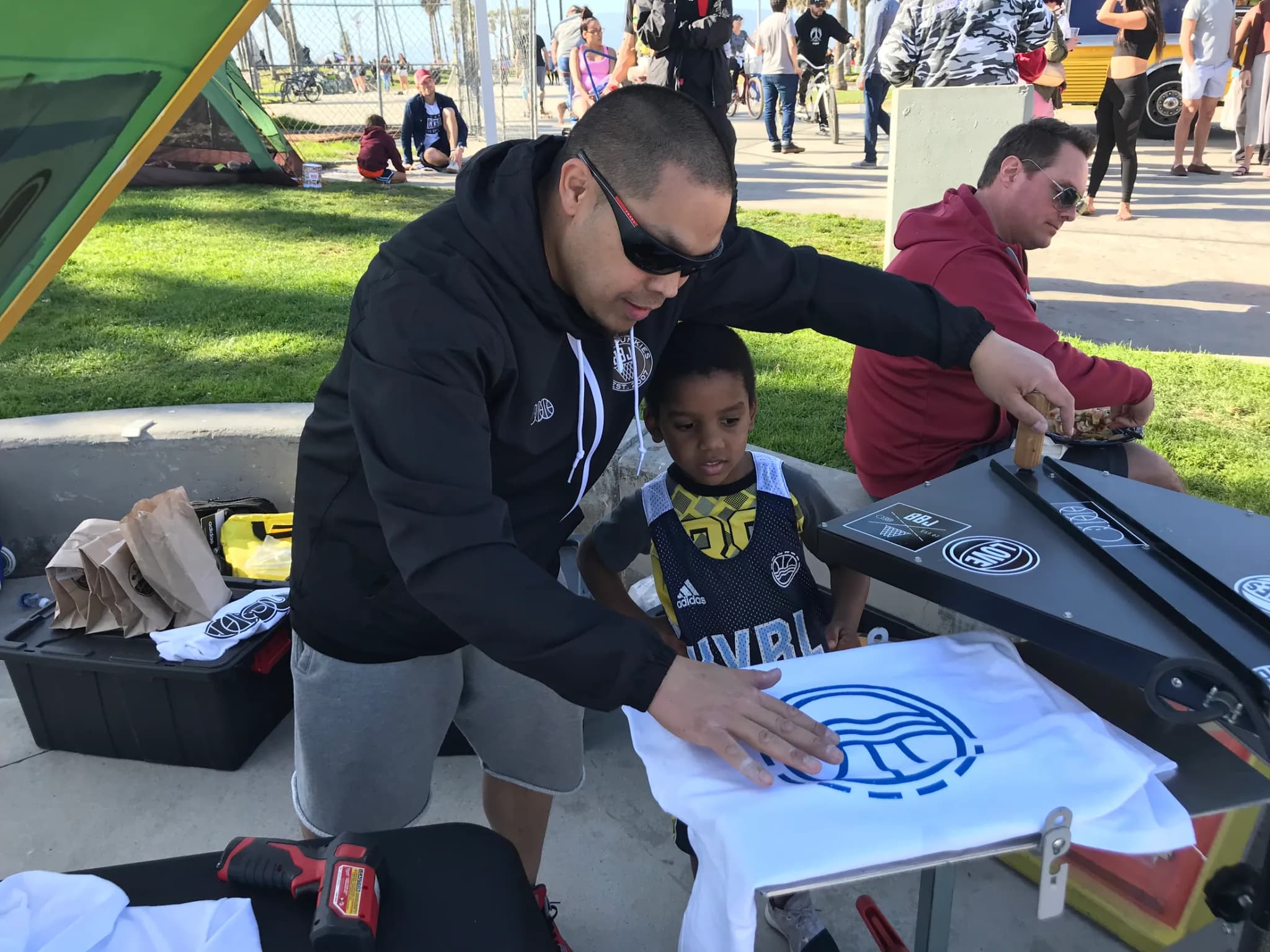 A printer in sunglasses smoothing a freshly printed white tee on a court-side press, the BBJ icon still wet at the chest, a young boy in a league jersey watching with his hand at the platen and the park crowd behind.