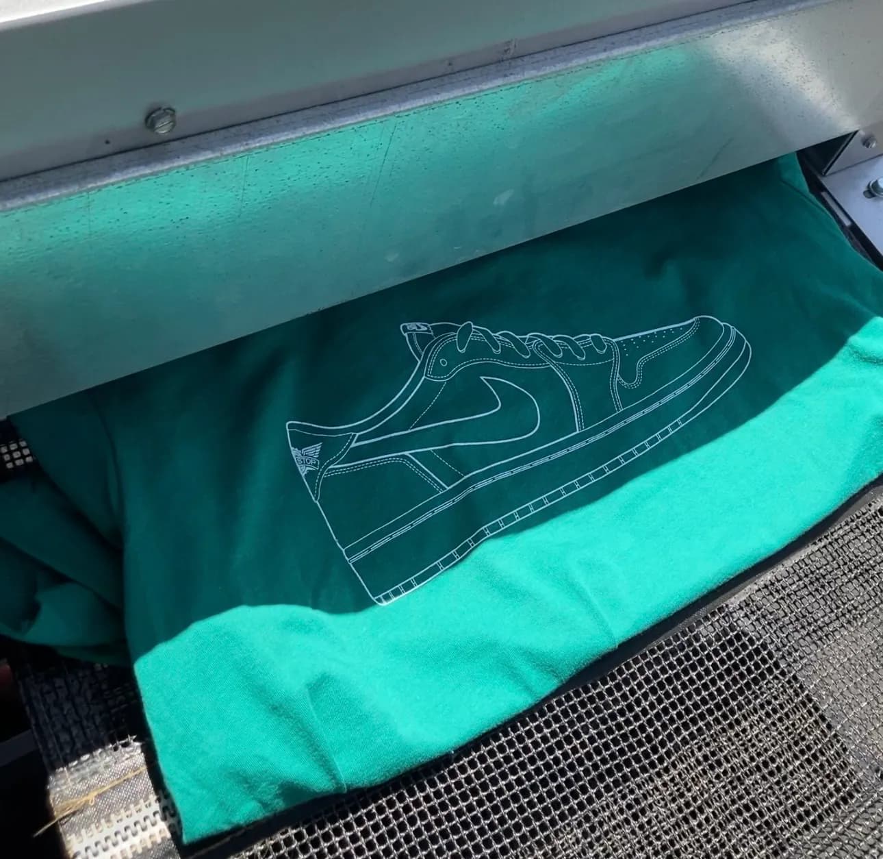 A Jordan 1 drawn in a single white outline across the chest of a mint green tee, riding the mesh belt out of the conveyor dryer.