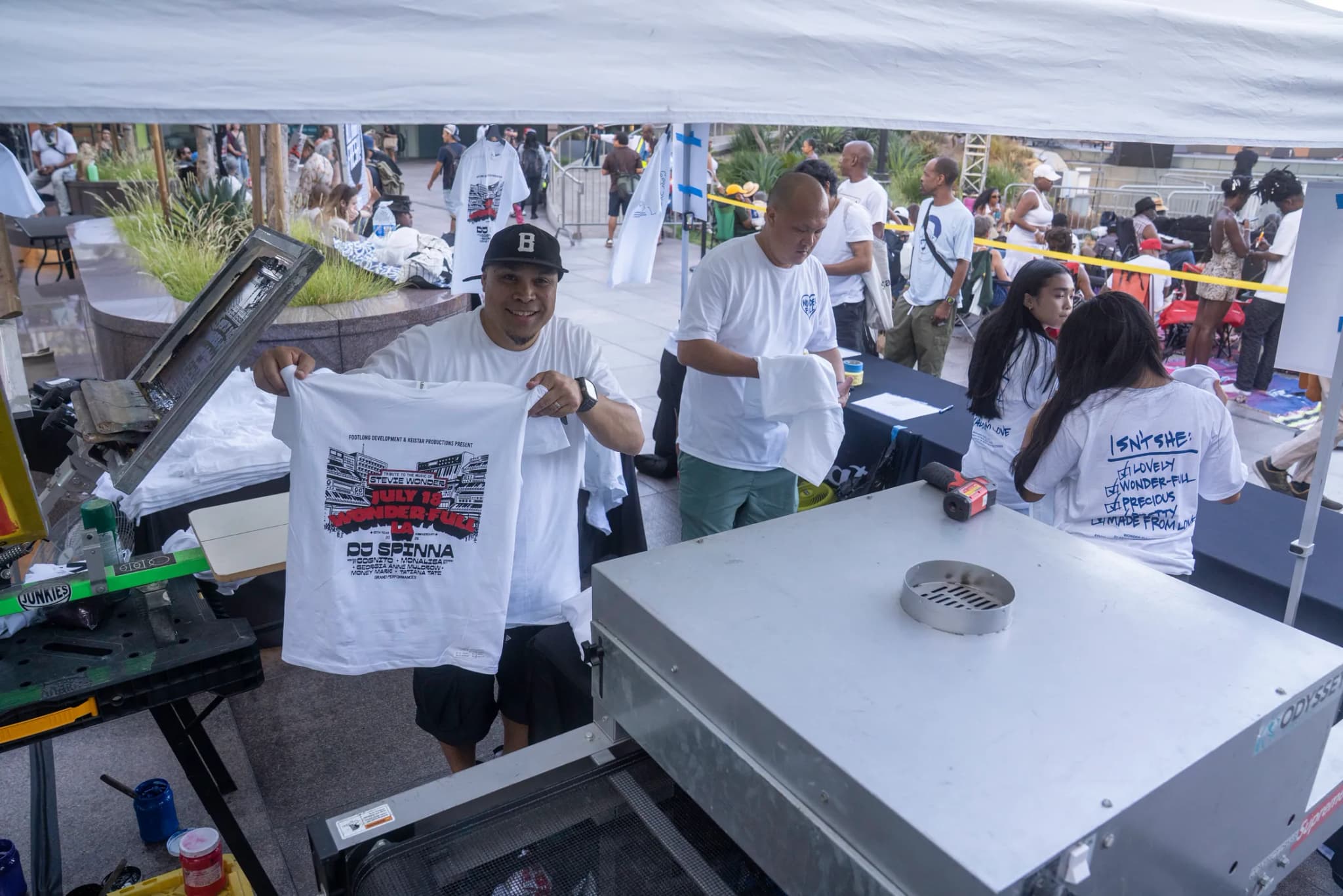 A printer in a black cap holding up a freshly printed white Wonder-Full LA tee under a white event tent, the conveyor dryer in front of him and a line of guests waiting along the taped-off table behind.