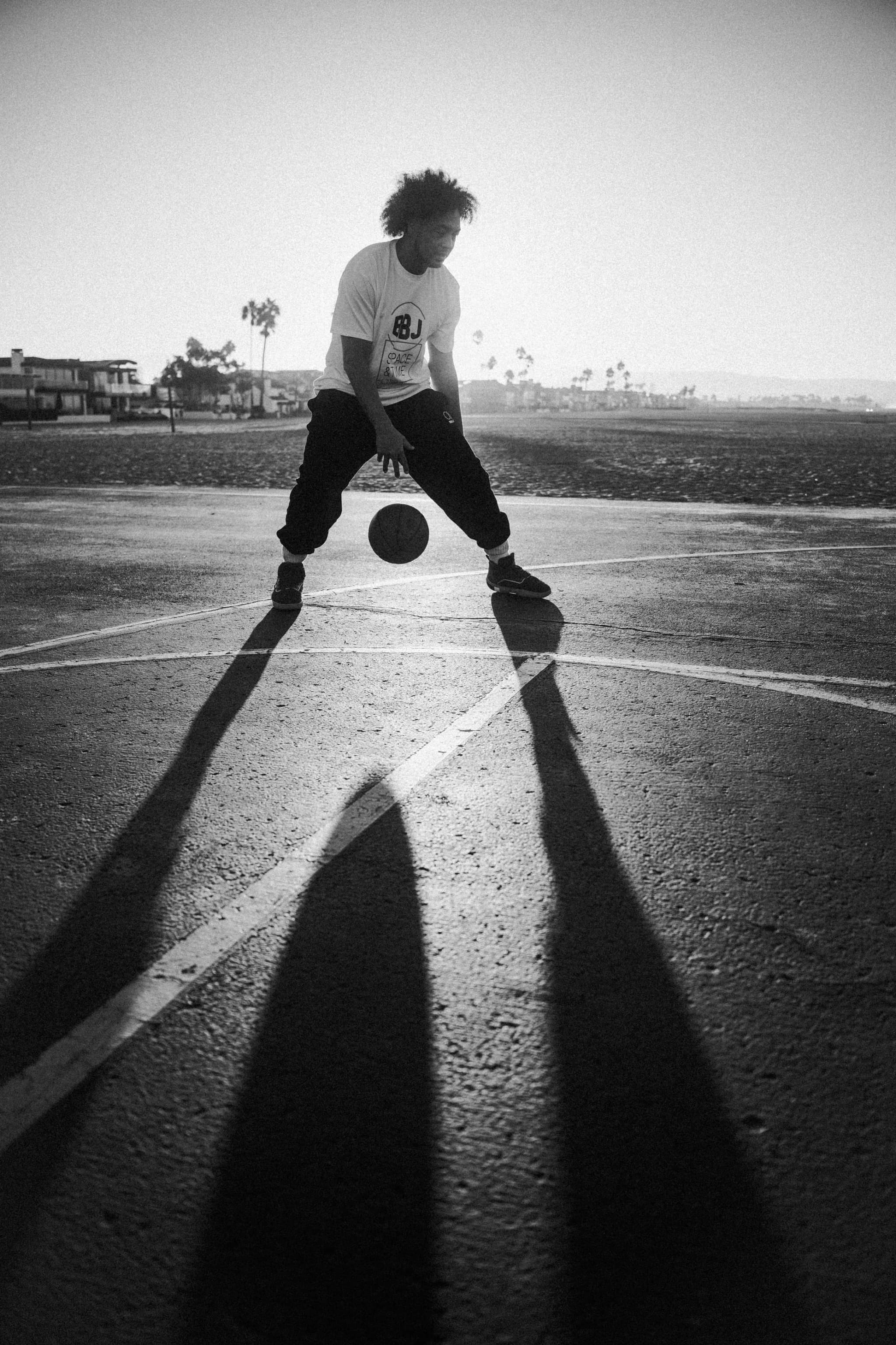 A player dribbling low on a beachside court in a white BBJ tee and dark pants, his long shadow thrown across the painted lines.