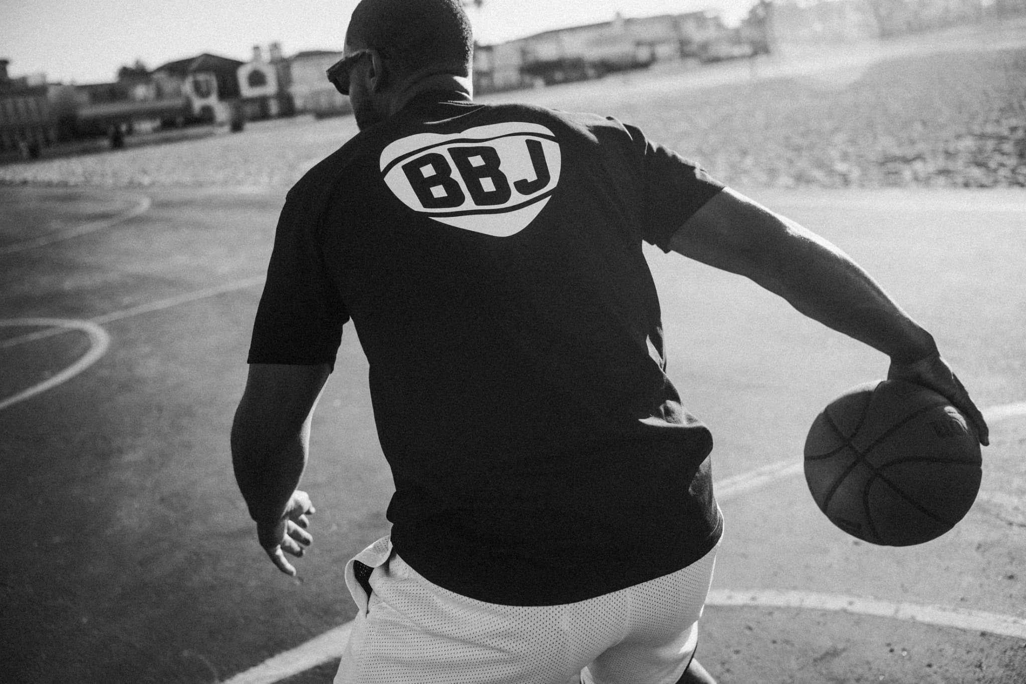 A player seen from behind dribbling low across a beach court, the BBJ heart mark across the back of his black tee, white shorts, the boardwalk soft beyond