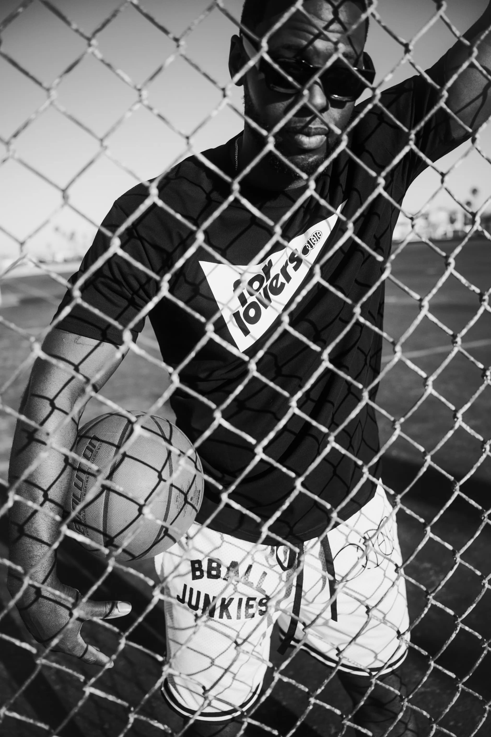 A player seen through a chain-link fence in sunglasses, the black For Lovers tee and white BBALL JUNKIES shorts.