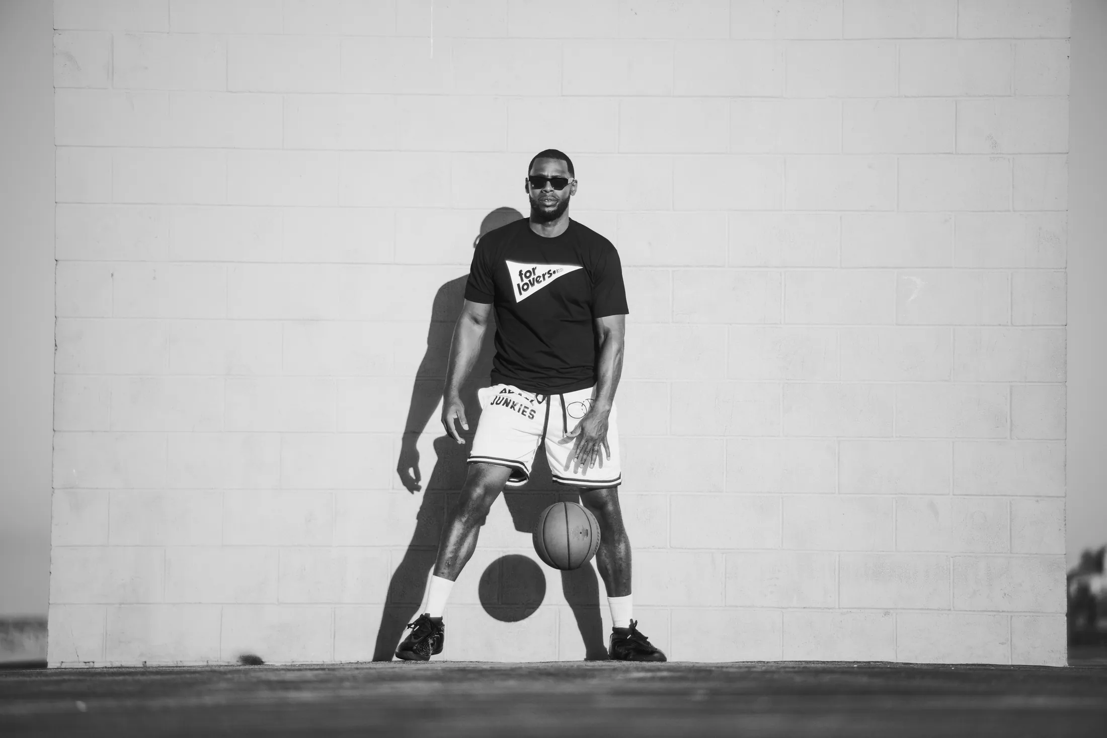 A player squared up in sunglasses against a white cinder-block wall, the ball between his feet, in a black For Lovers tee and white BBALL JUNKIES shorts, his shadow thrown onto the block
