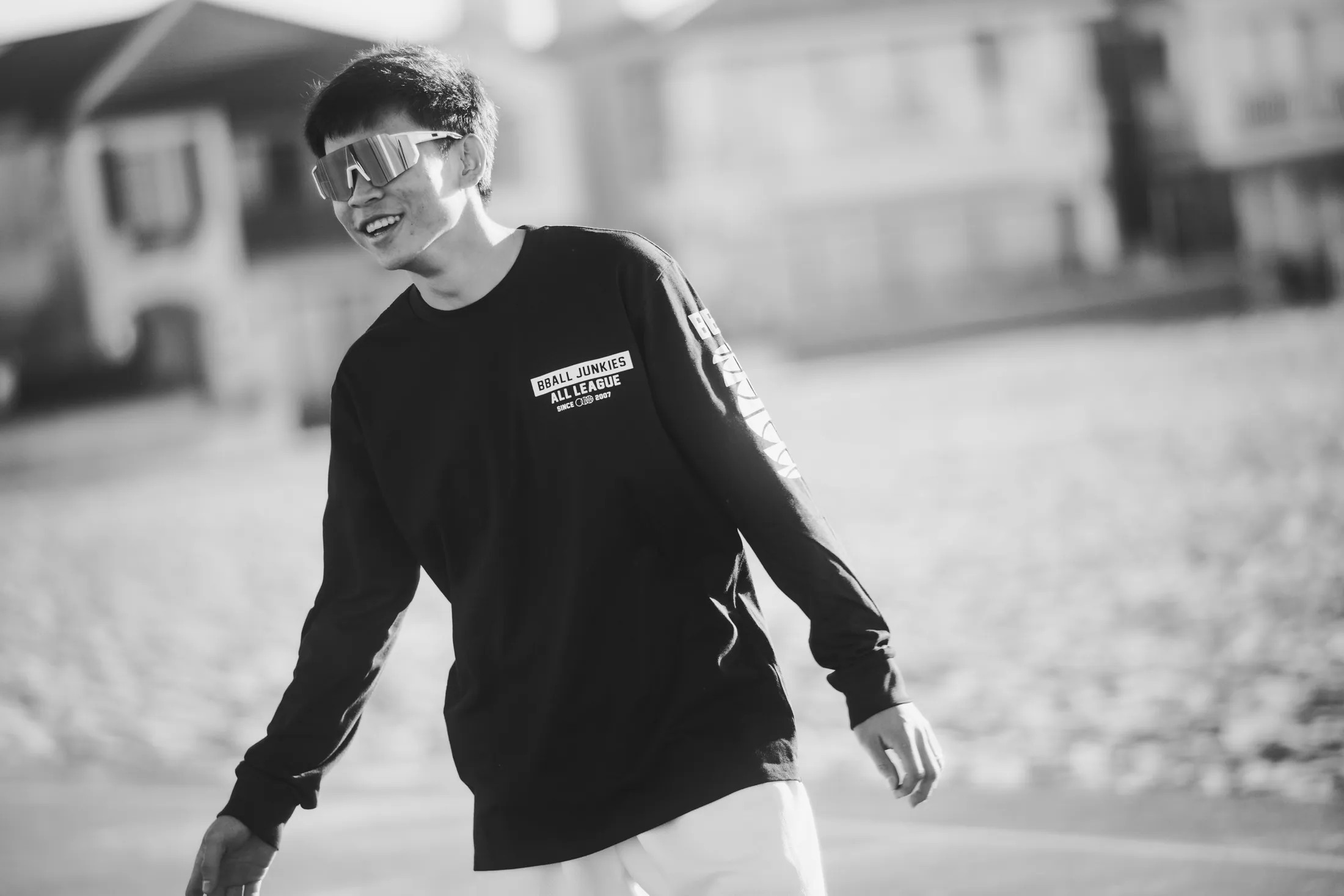 A player in mirrored shades laughing mid-dribble in the black All League long sleeve, beach houses out of focus behind him.