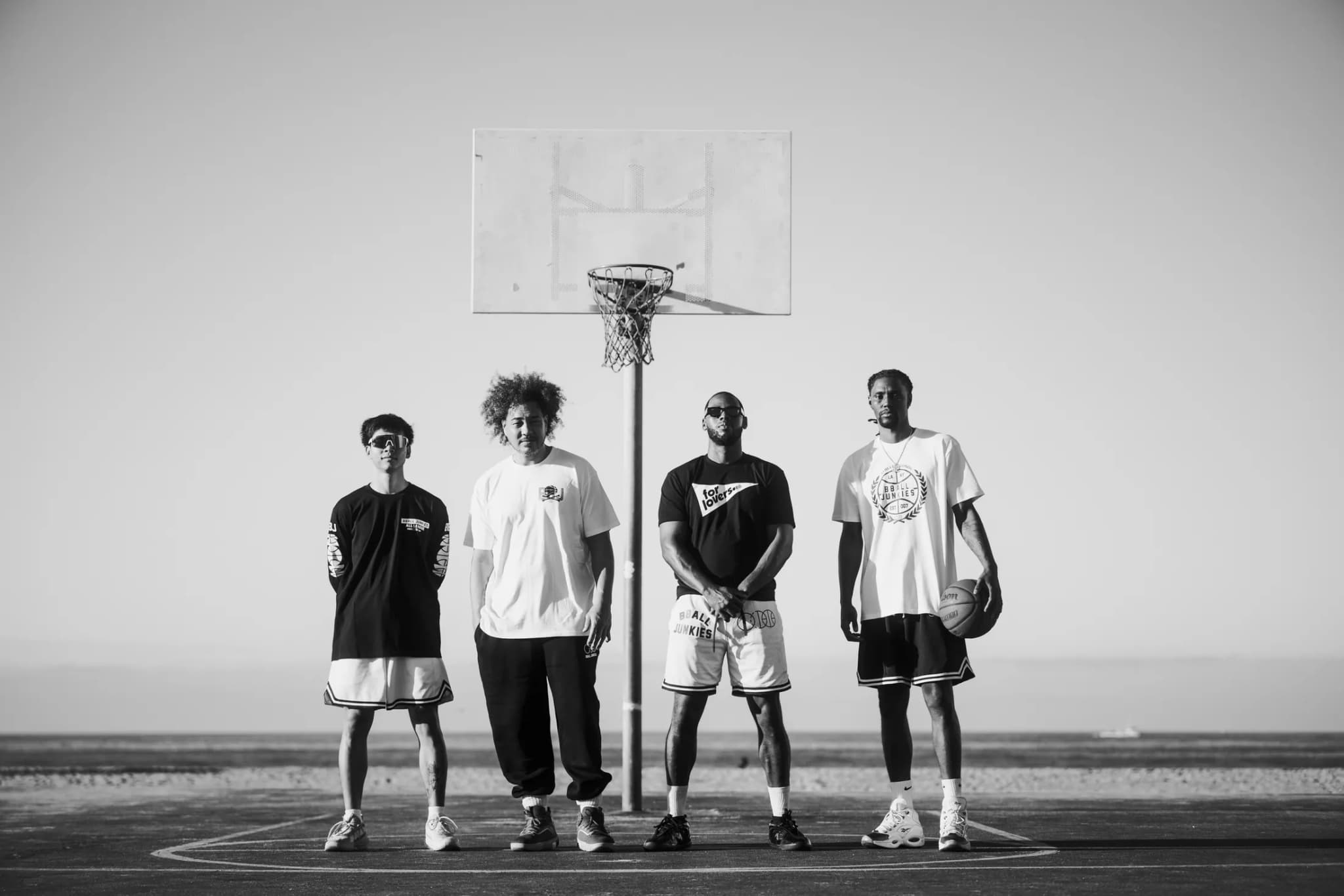 Four players lined up under a beachside hoop in BBJ tees and shorts, the ocean flat behind them.
