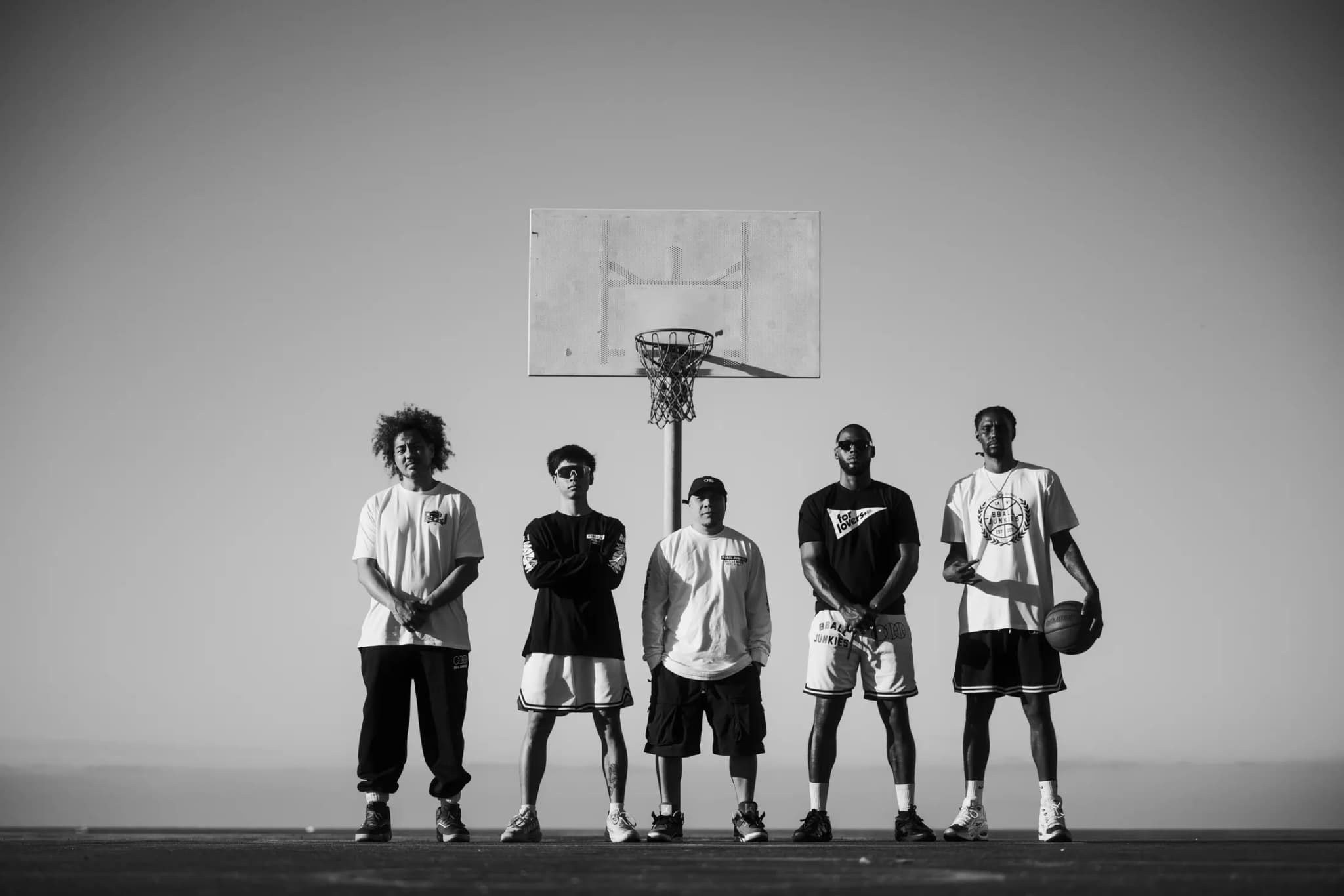 Five players lined up under a beachside hoop in BBJ tees, crewnecks and shorts, one holding a ball.