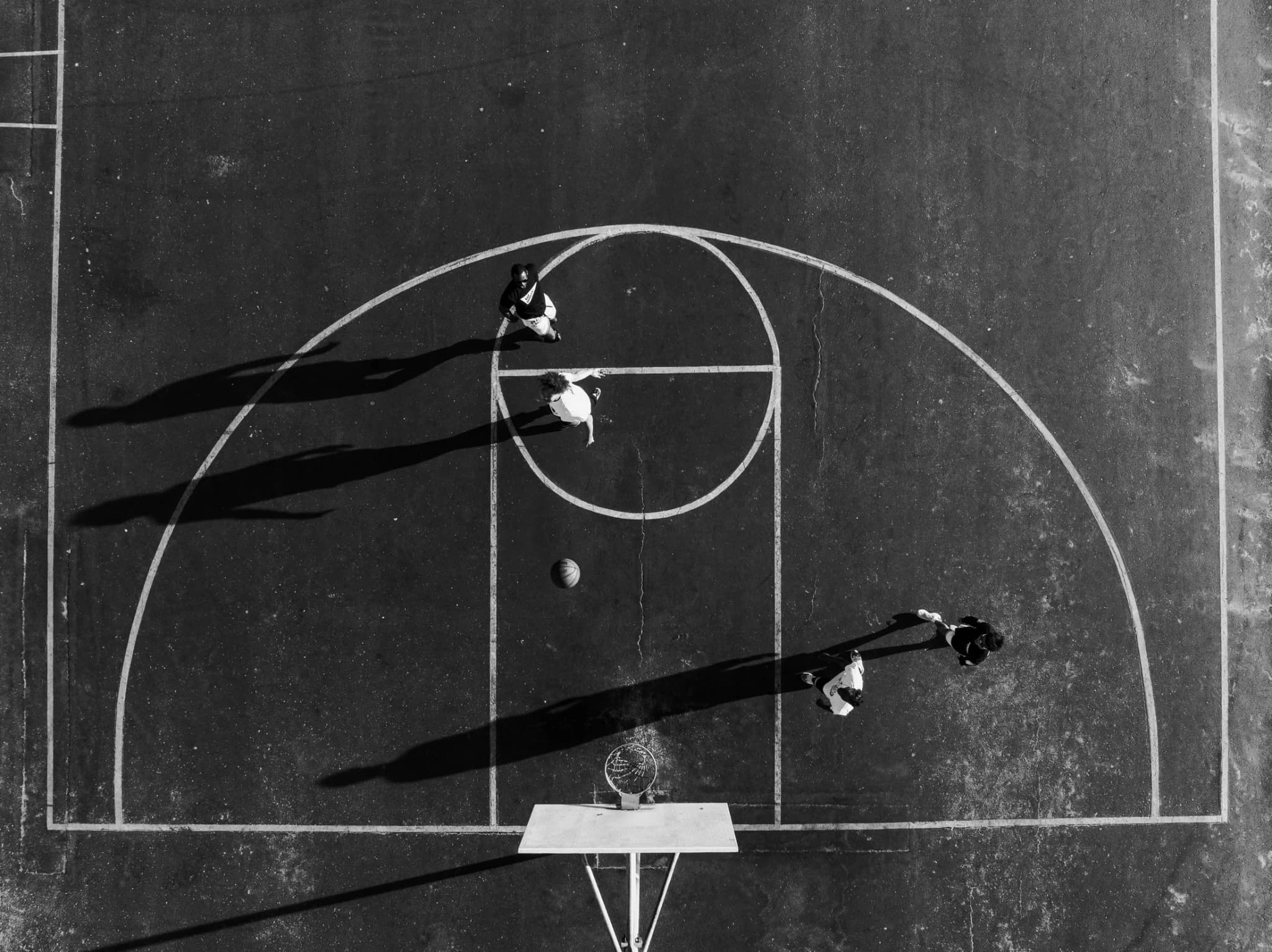 The court from directly overhead: two players and a loose ball on the painted key, their shadows running long across the asphalt.