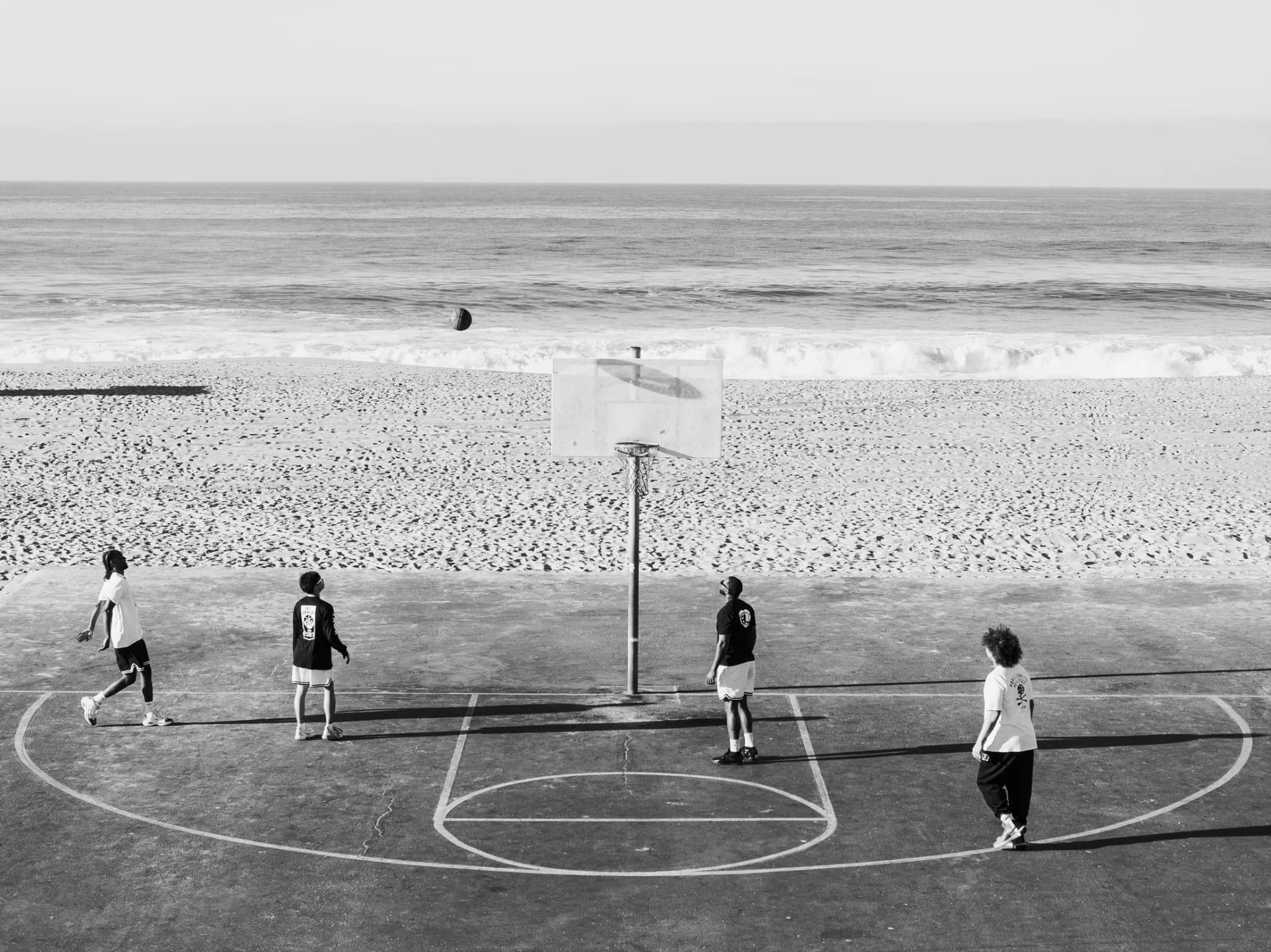 A beachside court from above, four players spaced along the baseline under a single hoop, the ocean behind.
