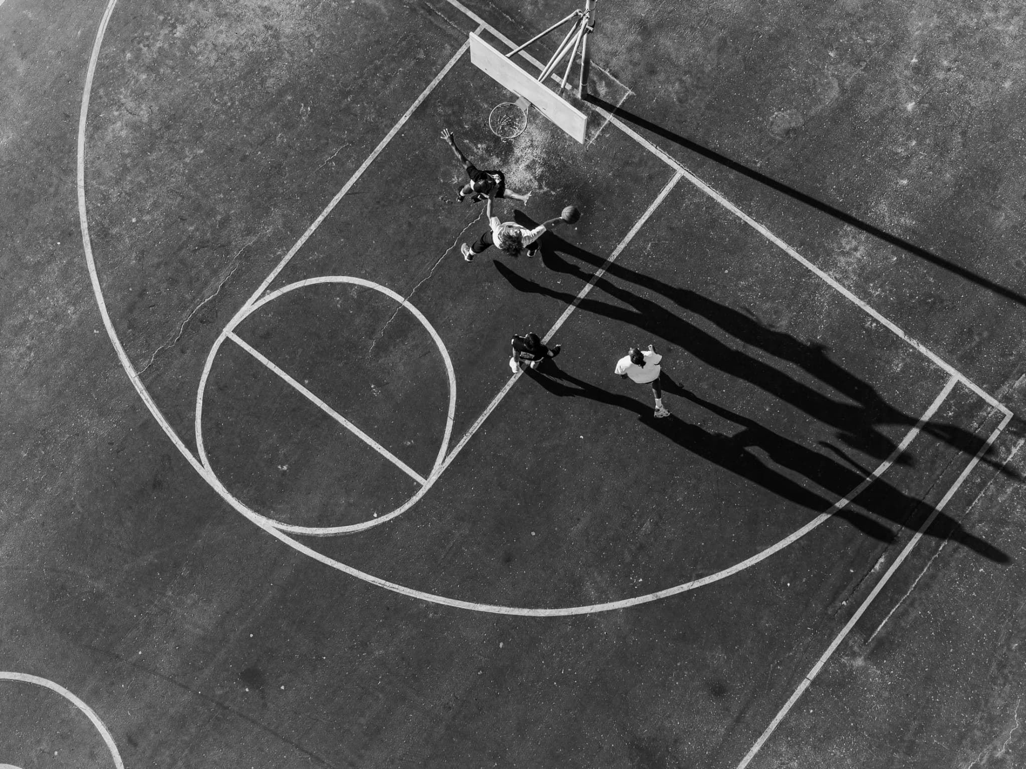 The court from overhead at an angle: three players and their long shadows inside the painted three-point arc.