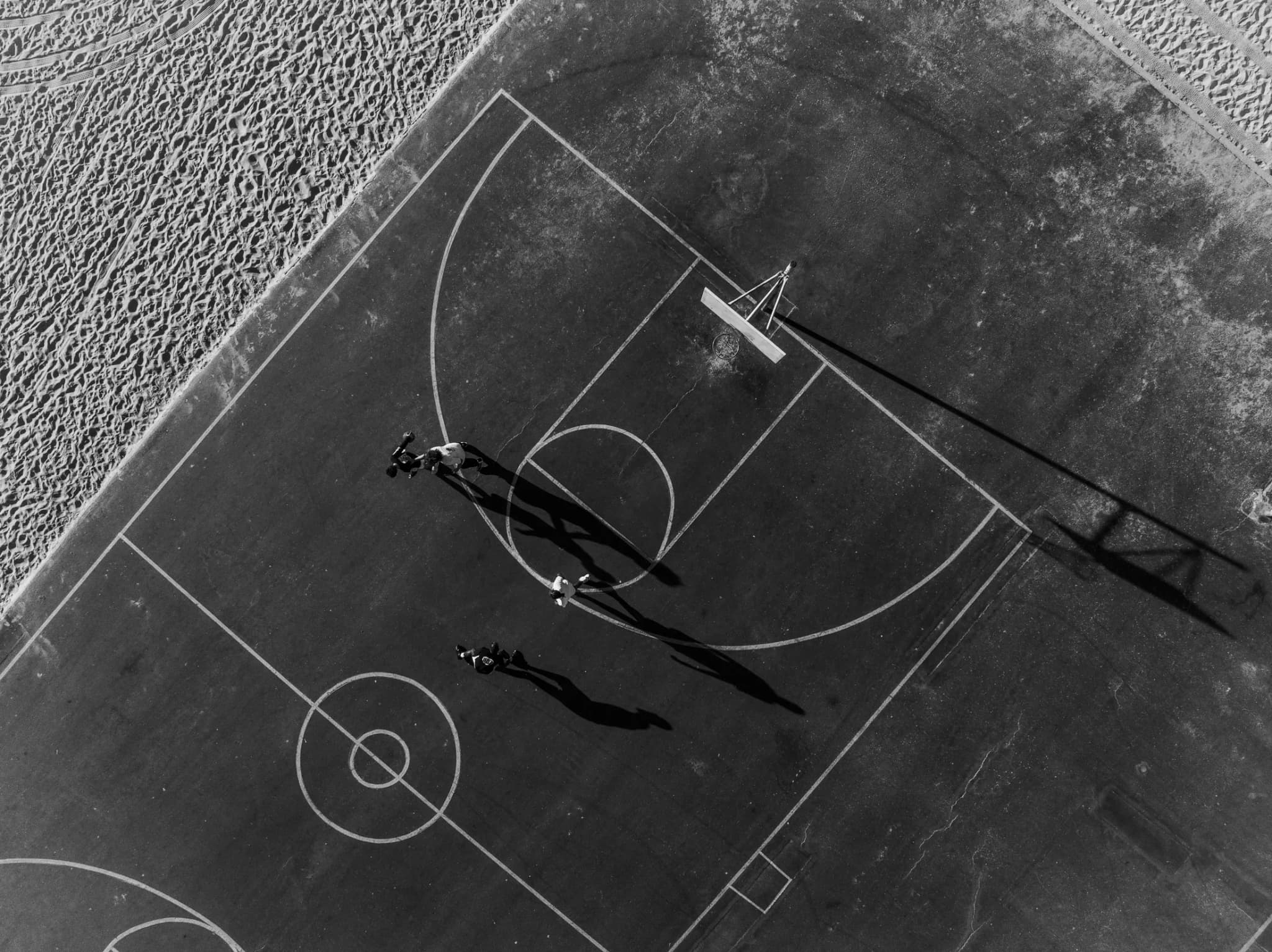 A beachside court from directly overhead, three players and their long shadows across the painted key, the sand at the court’s edge.