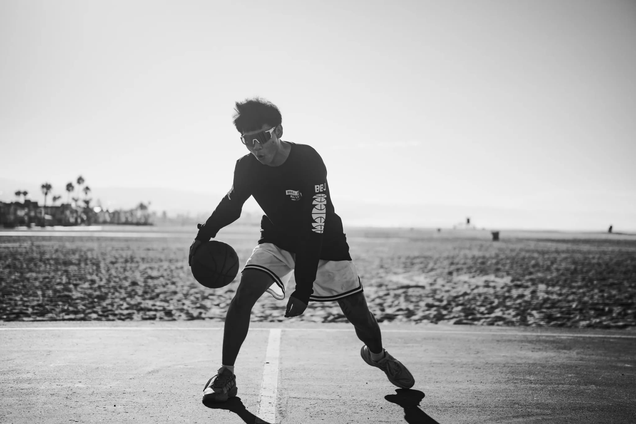 A player in mirrored shades crossing the ball over low, in a black All League long sleeve and white shorts, the beach behind him.